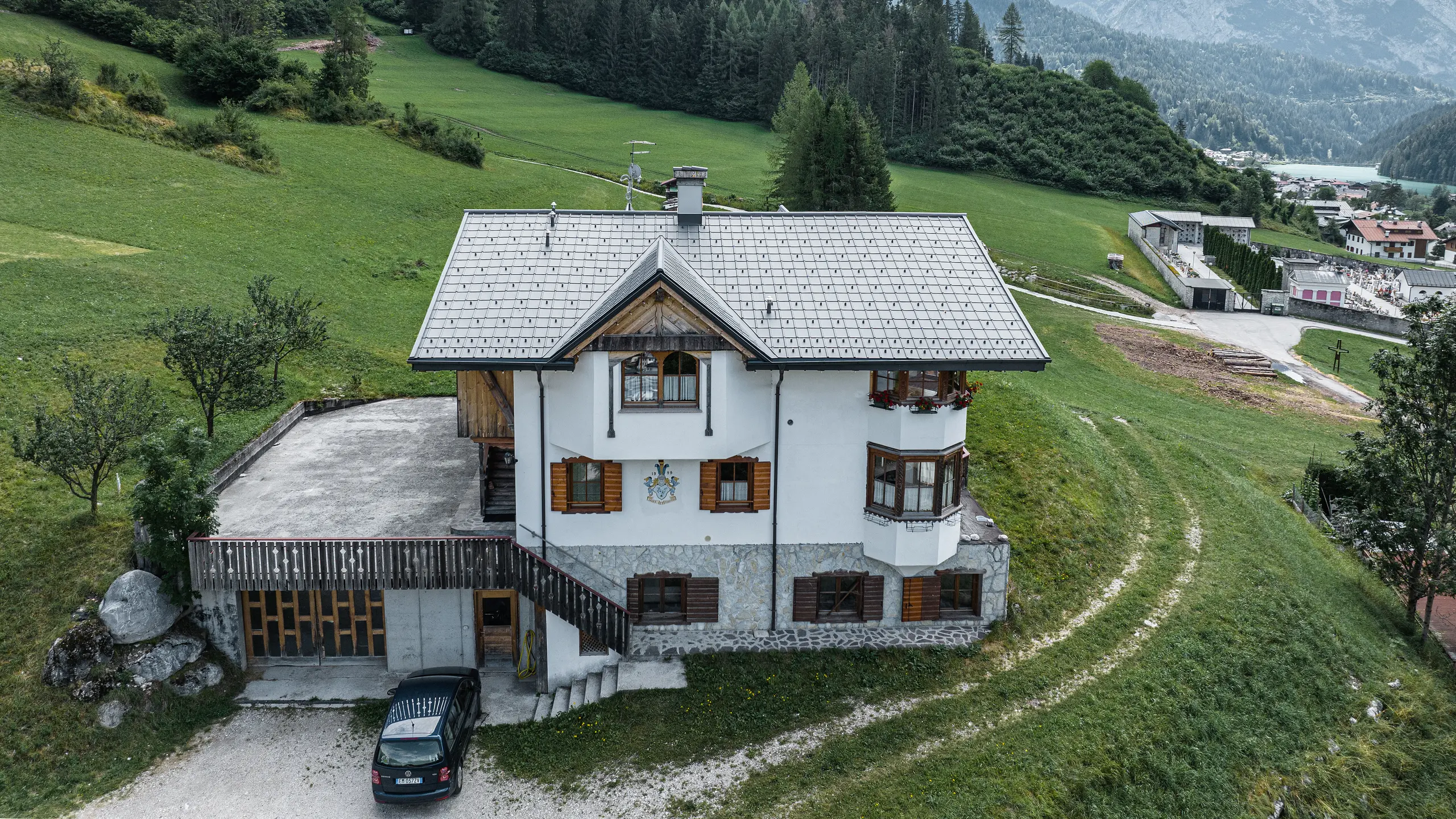 Pohled z ptačí perspektivy na vchod do tradiční horské chaty v Auronzo di Cadore po rekonstrukci střechy, vyznačující se falcovanými taškami PREFA v barvě P.10 světle šedé. Budova s ​​charakteristickým alpským šarmem a moderní střechou je v harmonii se svěží zelenou krajinou a působivým horským panoramatem v pozadí a demonstruje úspěšnou kombinaci funkčnosti a regionální architektury.