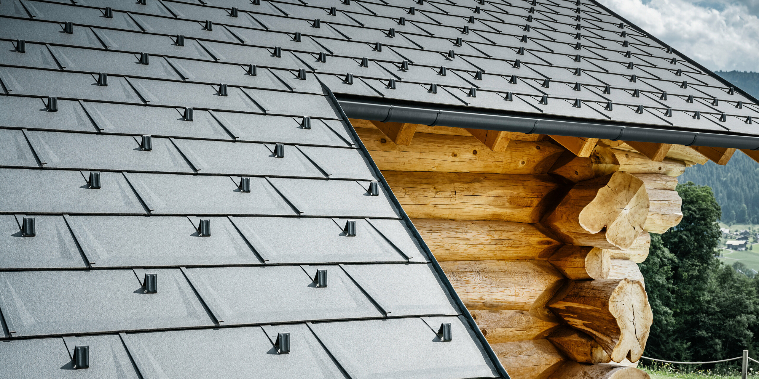 Detailní pohled na střechu se střešními panely PREFA R.16 v barvě P.10 antracitové na srubovém domě v Gosau, Horní Rakousko. Precizní zpracování a montáž robustních střešních panelů dokazuje vysokou kvalitu hliníkových výrobků PREFA a také dokonalou integraci moderních hliníkových komponentů do tradičních typů budov z Rakouska. V popředí jsou vidět charakteristické sněhové zábrany, které zajišťují bezpečnost v alpské zimě. Tento obrázek zachycuje harmonickou interakci inovativních střešních systémů PREFA s přirozenou estetikou dřevěného srubu.