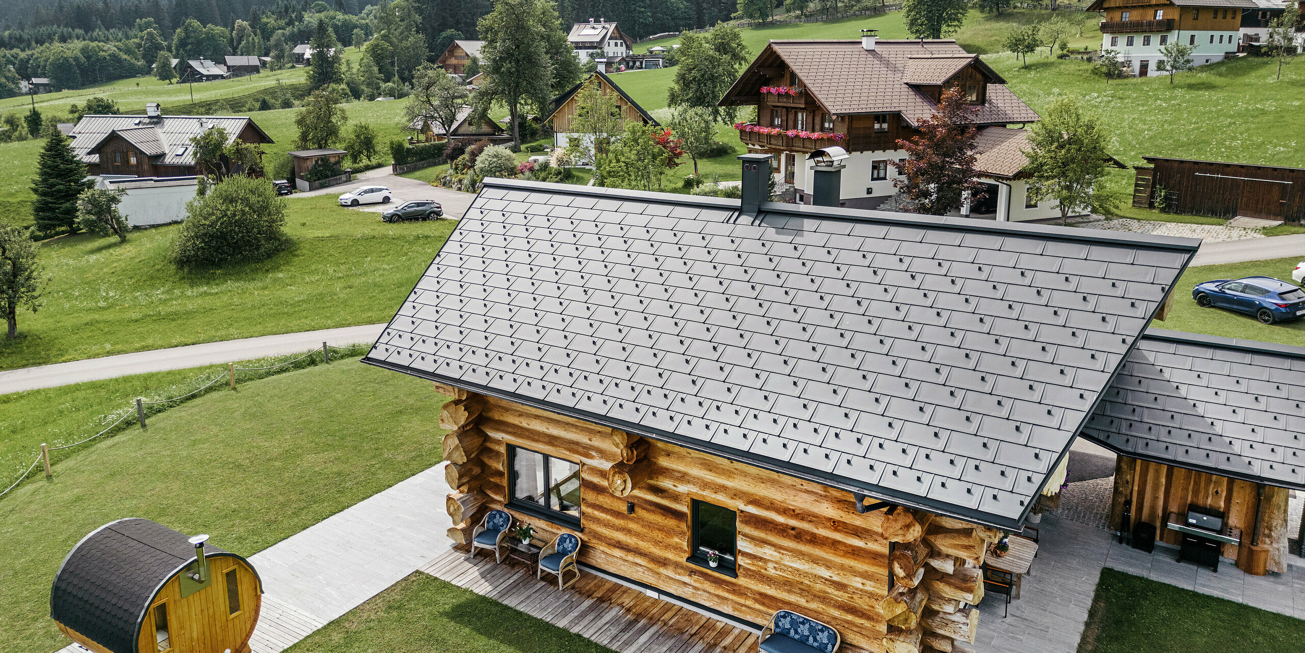 Pohled z ptačí perspektivy na rustikální srub v rakouském Gosau s kvalitní hliníkovou střechou PREFA ze střešních panelů R.16 v barvě P.10 antracitové. Střecha ukazuje typickou strukturu a preciznost střešních systémů PREFA, které dokonale zapadají do venkovského prostředí. Svěží zeleň luk, okolní tradiční domy a zalesněné kopce v pozadí tvoří přirozený kontrast k moderním střešním prvkům v antracitové barvě, které do alpské architektury přinášejí odolnost a moderní estetiku.