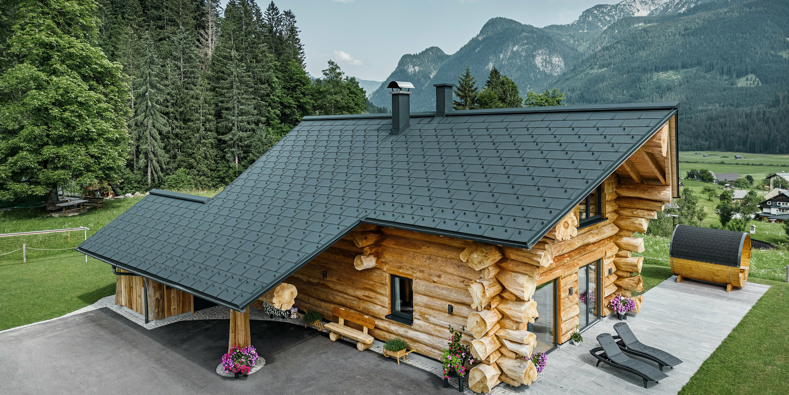 Šikmý pohled na malebný srub v rakouském Gosau vybavený střechou PREFA ze střešních panelů antracitové barvy R.16. Architektura kombinuje styl rustikálního srubu s moderními prvky, obklopený hustými zelenými lesy a působivou horskou kulisou. Dům vyzařuje přívětivou atmosférou svou venkovskou elegancí a precizně položenými hliníkovými střešními prvky, které jsou známé svou životností a odolností vůči povětrnostním vlivům.