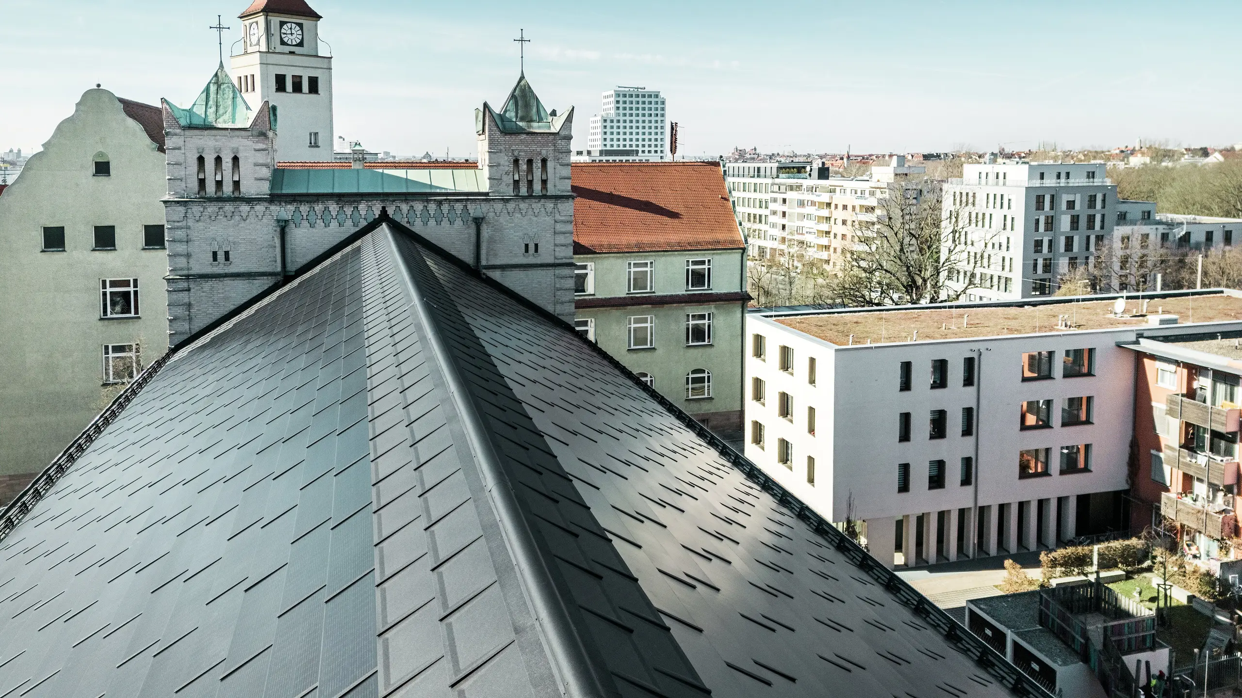 Das Bild zeigt die präzise Deckung des Aluminiumdachs der St. Karl Borromäus Kirche in Nürnberg. Das Kirchendach wurde mit PREFA Solardachplatten in elegantem Schwarz ausgestattet. Der Blick auf den markanten First des Satteldachs unterstreicht die gelungene Integration der modernen Photovoltaik-Indach-Lösung in die historische Architektur. Die Solardachplatten fügen sich harmonisch auf dem denkmalgeschützten Gebäude ein und versorgen die Kirche und etliche benachbarte Gebäude mit effizienter und nachhaltiger Energie.