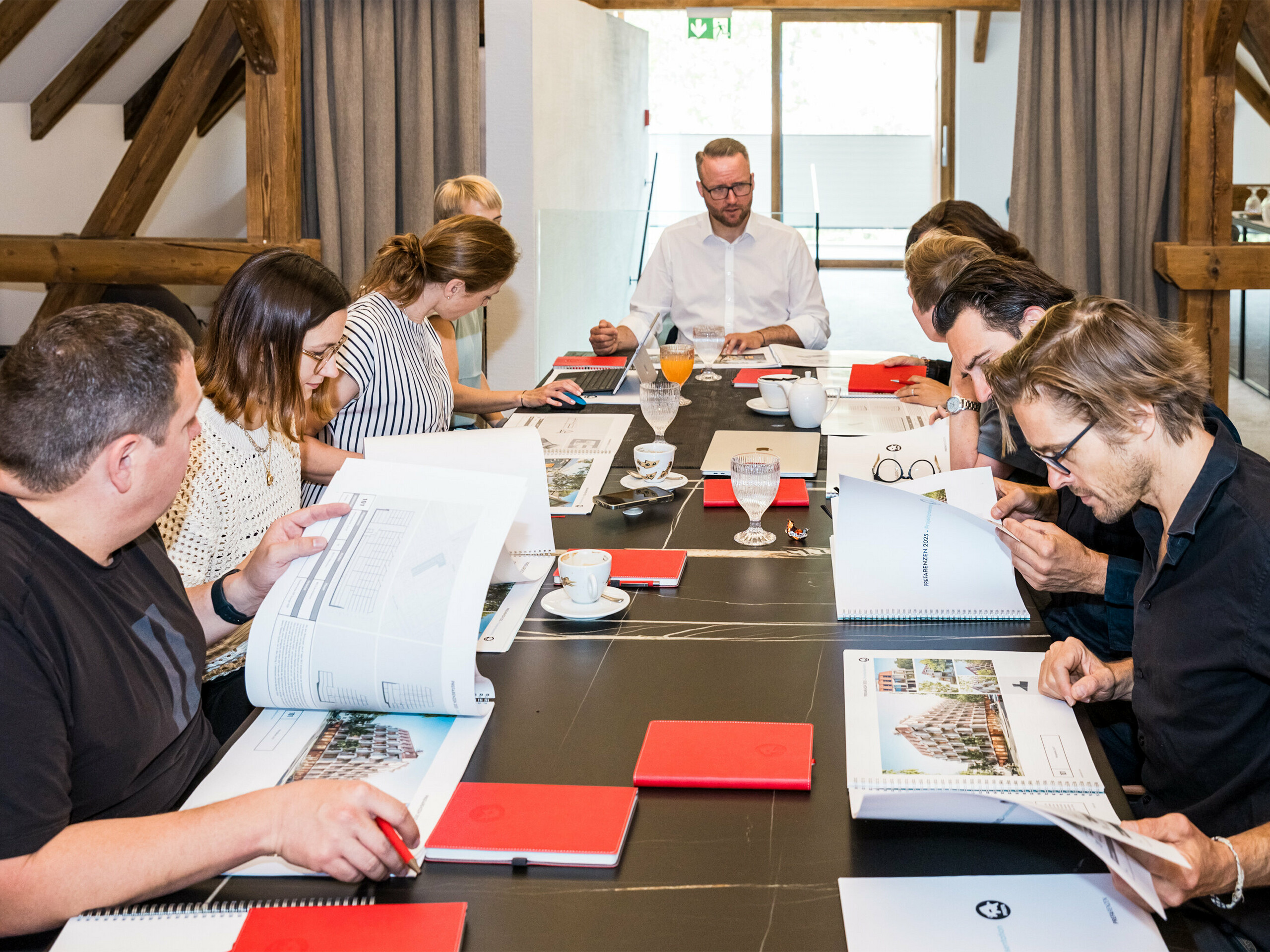 PREFARENZEN 2025 Odborný dialog: Deset architektů a expertů PREFA sedí u konferenčního stolu v sídle vinaře v Seewinkelu v Burgenlandu. Na stole jsou dokumenty a architektonické plány, zatímco účastníci intenzivně diskutují a vybírají dvanáct nejzajímavějších architektonických objektů pro PREFA Architecture Platform 2025. Diskuse je věnována ekologickým a společenským výzvám, funkčnosti, jednoduchosti a přidané hodnotě architektury. Na stole jsou také rozházené kávové šálky, sklenice a červené sešity, což podtrhuje soustředěnou pracovní atmosféru.