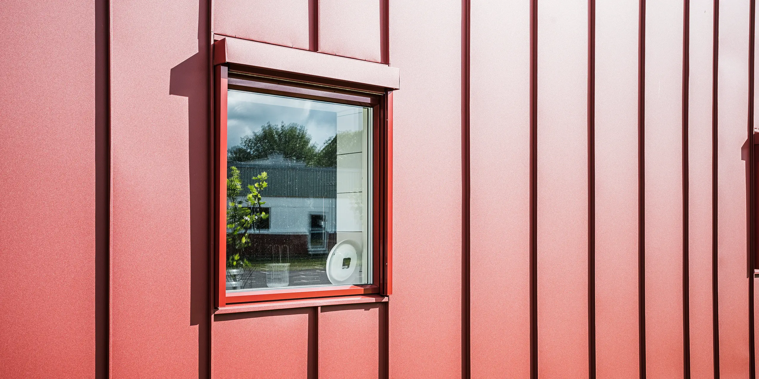 Detailní záběr na okno školní jídelny Gröna Hjärtat ve švédském Broby, zobrazující detailní červenou fasádu z PREFALZ. Precizní zpracování hliníkové střešní krytiny se stojatou drážkou podtrhuje vysokou kvalitu a dlouhou životnost pláště budovy. Okno je harmonicky začleněno do fasády a skleněná výplň odráží okolí. Atraktivní design činí z budovy příklad moderní a udržitelné výstavby.