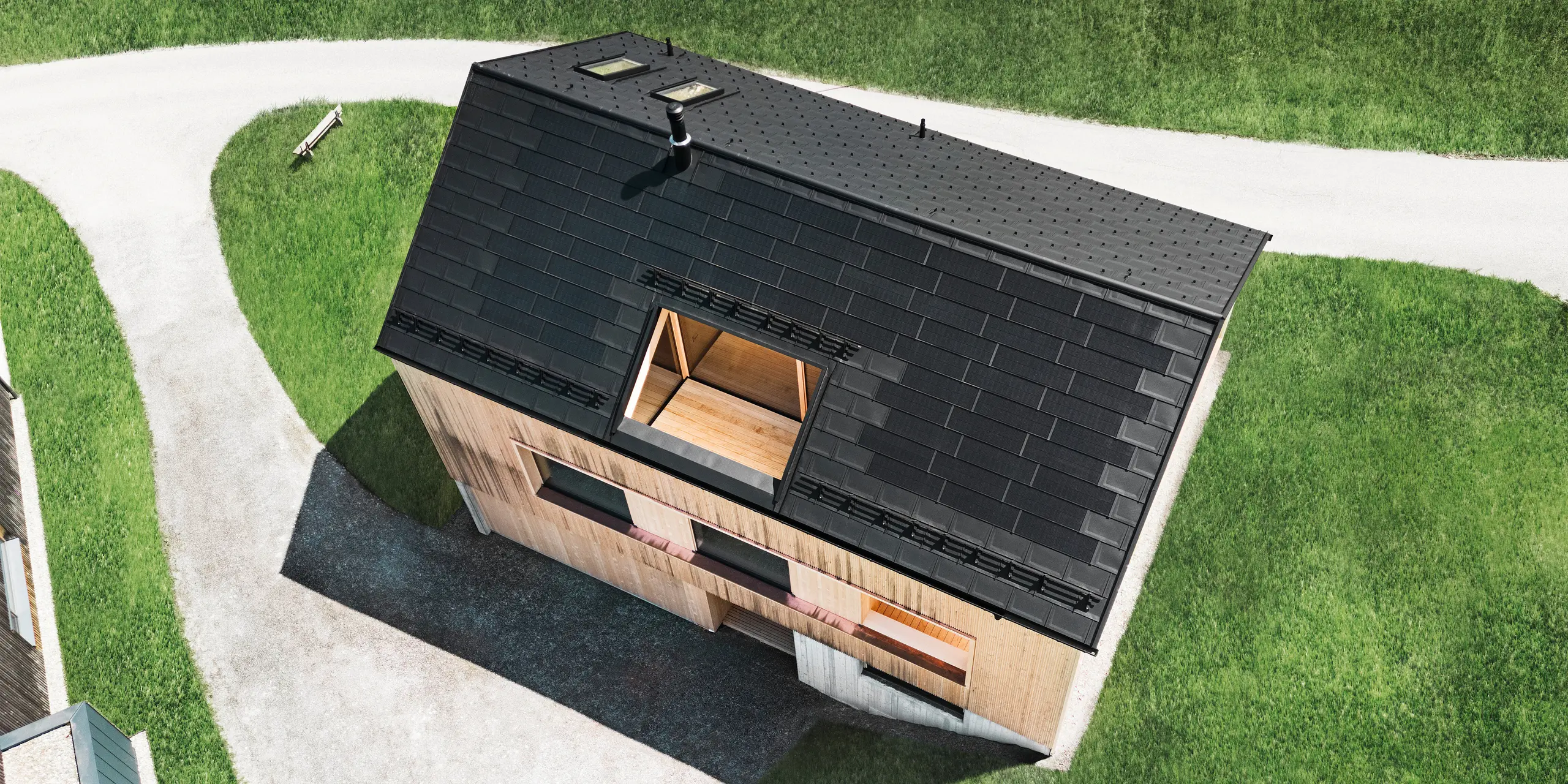 Záběr z dronu s pohledem shora na ekologický rodinný dům ve Eggu, který vyčnívá z okolních zelených trávníků černou solární střechou PREFA a tradičními střešními panely PREFA R.16. Strategicky umístěné solární moduly maximalizují energetickou účinnost, zatímco jasná geometrie střechy a přirozeně vypadající dřevěná fasáda vytvářejí spojení mezi moderním bydlením a venkovským prostředím. Tento dům ztělesňuje progresivní design šetrný k životnímu prostředí, který zapůsobí jak funkčně, tak vizuálně.