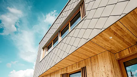 Detailní záběr na školku v Alzenau ukazuje precizní zpracování a vysoce kvalitní kombinaci bronzově zbarvených stěnových panelů PREFA (44 x 44 cm) a přírodního dřeva. Diagonální struktura hliníkové fasády vytváří výrazný šupinovitý vzor, ​​který krásně ladí s vertikálními dřevěnými žaluziemi kolem oken a precizně zpracovaným dřevěným podhledem. Záběr z nízkého úhlu zdůrazňuje přesné linie a řemeslné zpracování odvětrávaného fasádního obkladu. Tento detailní pohled podtrhuje konstrukční pevnost a trvanlivost produktů PREFA – ideálních pro udržitelné architektonické projekty, které kombinují estetiku, funkčnost a ekologickou odpovědnost na nejvyšší úrovni.