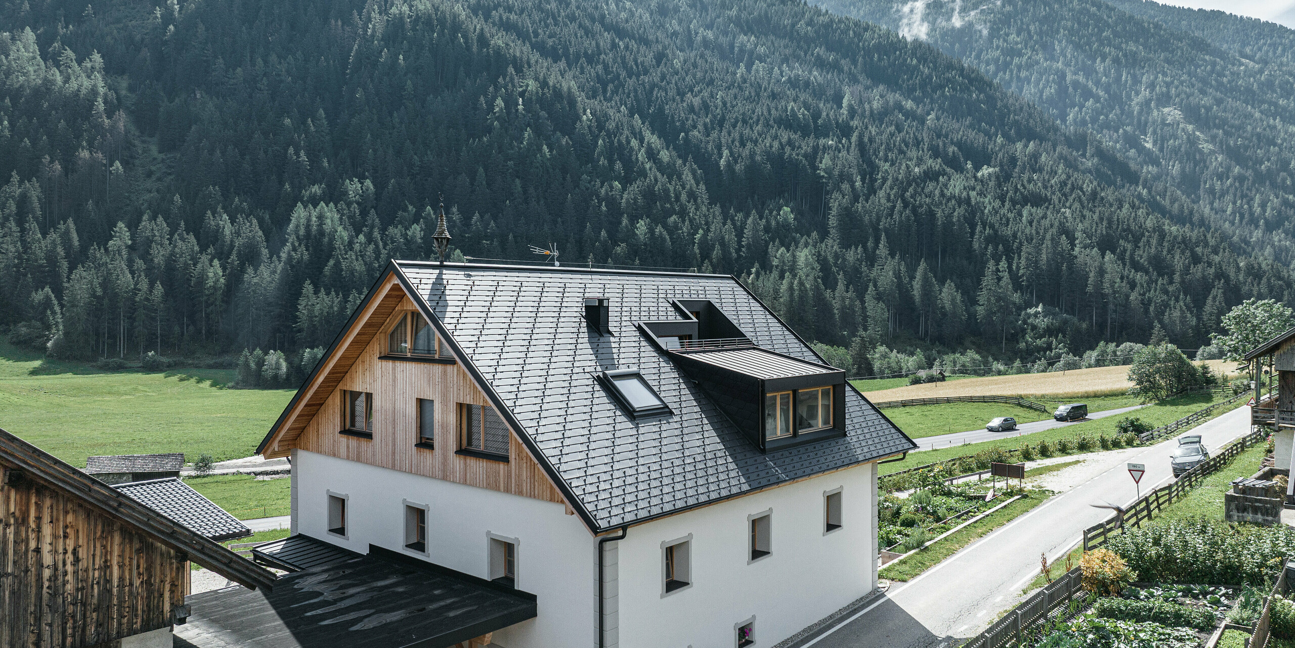 Perspektivní pohled na Ausserwegerhof v Antholz, Jižní Tyrolsko, s moderní střechou PREFA z šindele antracitové barvy DS.19. Bílá fasáda budovy vyniká proti hustému jehličnatému lesu a klidnému výhledu do ulice. Štíty budovy jsou obložené dřevem a poskytují harmonický přechod na hliníkovou střechu. Ze sedlové střechy na obou stranách vyčnívá vikýř. Pod vikýři jsou velkorysá okna, ze kterých je dobrý výhled.