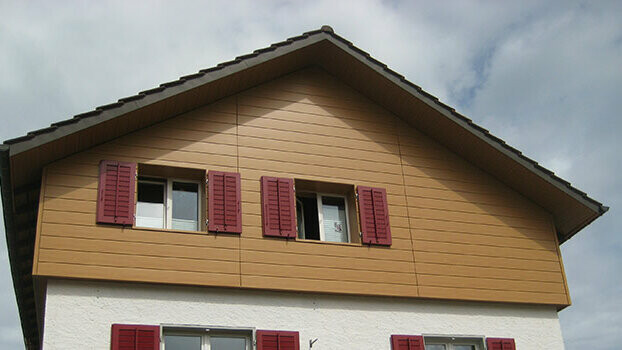 Obložení štítu klasického domu se sedlovou střechou. Štít je obložen horizontálně montovanými PREFA Sidings v designu dřeva (dub přírodní). Okna mají červené okenice.