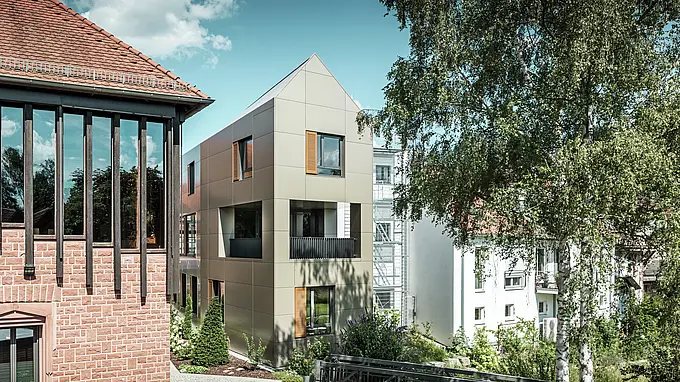 Na obrázku je moderní radnice v Alzenau, jejíž fasáda je vyrobena z bronzových hliníkových kompozitních panelů od společnosti PREFA. Špičatá střecha a jasně strukturovaná okna s dřevěnými žaluziemi dodávají budově elegantní a moderní estetiku. V popředí je vidět zelená plocha s dlážděnou cestou a stromy, které harmonicky doplňují urbanistický design budovy.