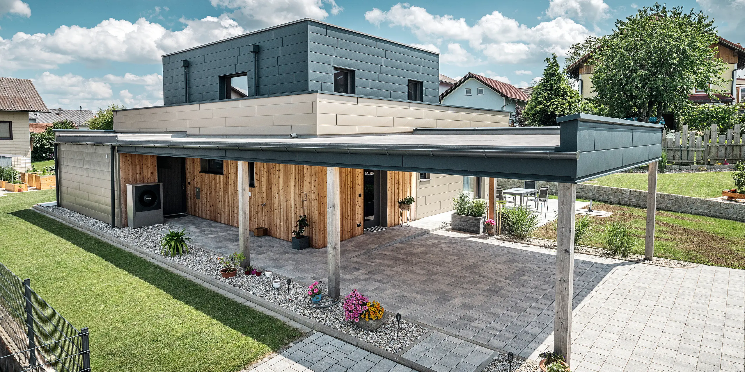 Letecký snímek příjezdové cesty k modernímu rodinnému domu ve Wippenhamu v Rakousku s dvoubarevnou hliníkovou fasádou PREFA v bronzové a antracitové barvě. Dům se vyznačuje stylovým začleněním fasádních panelů Siding.X a dřevěného obložení. Zřetelně viditelný je krytý přístřešek pro auto, který je hladce integrován do architektury domu. Upravená zahrada a terasa podtrhují moderní a funkční design budovy.