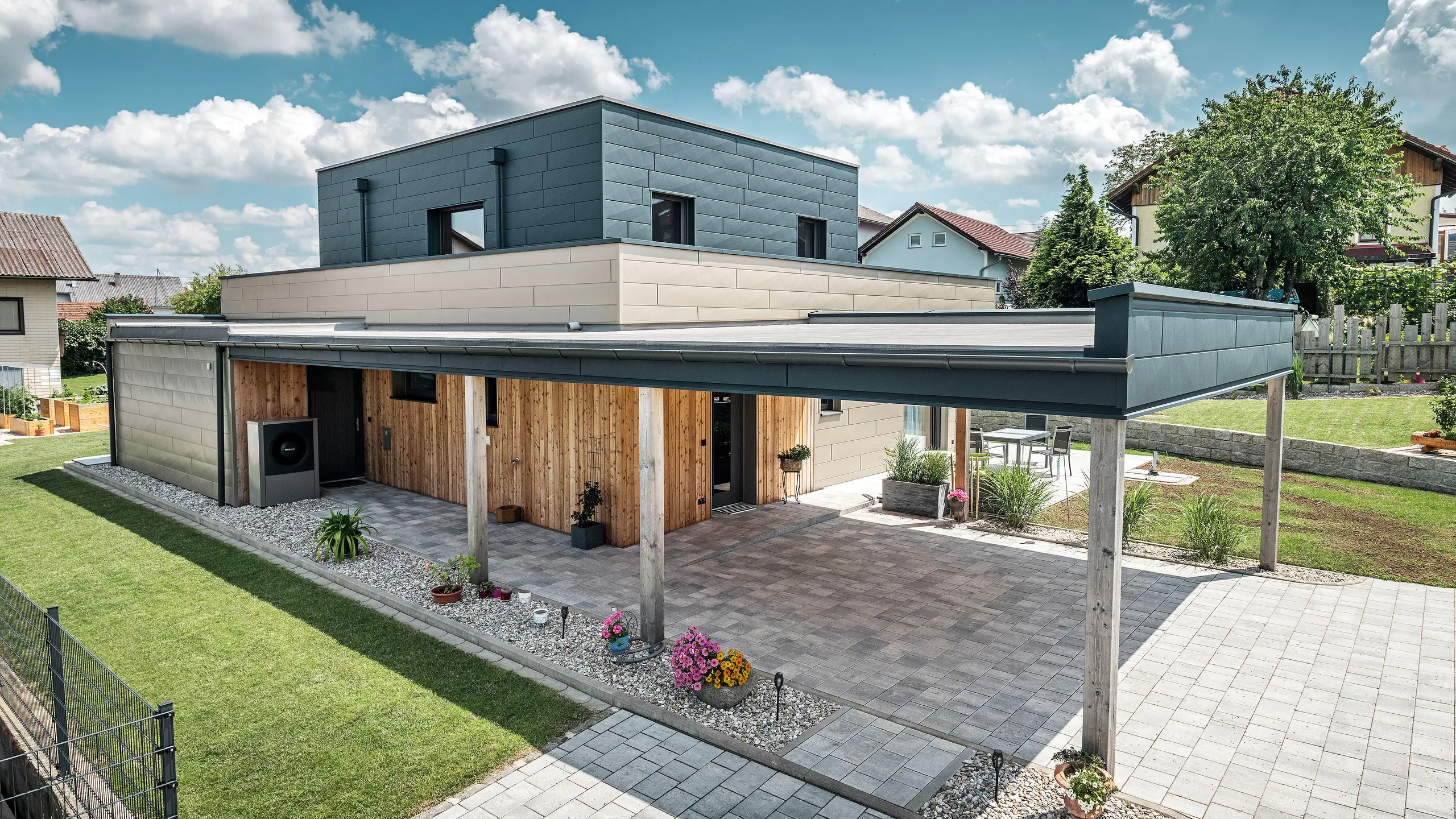 Letecký snímek příjezdové cesty k modernímu rodinnému domu ve Wippenhamu v Rakousku s dvoubarevnou hliníkovou fasádou PREFA v bronzové a antracitové barvě. Dům se vyznačuje stylovým začleněním fasádních panelů Siding.X a dřevěného obložení. Zřetelně viditelný je krytý přístřešek pro auto, který je hladce integrován do architektury domu. Upravená zahrada a terasa podtrhují moderní a funkční design budovy.