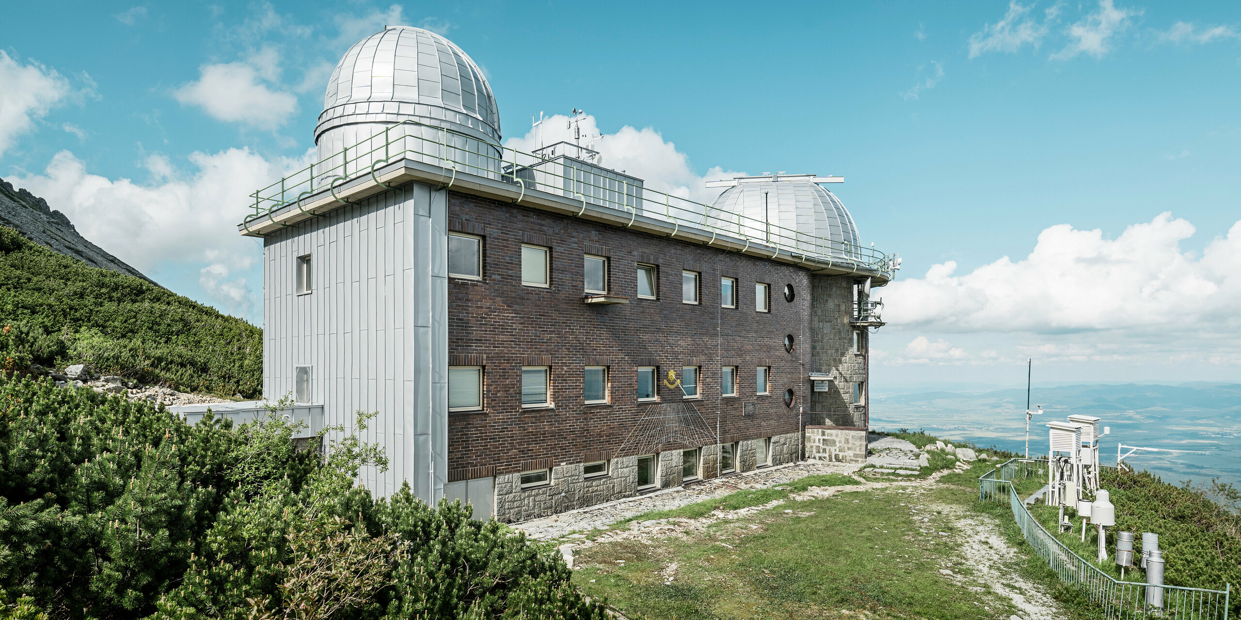 Boční pohled na hvězdárnu Skalnaté Pleso, harmonicky zasazenou do přírody s PREFALZ ve stříbrné metalíze a tradiční cihlovou fasádou. Observatoř, která se nachází ve slovenské horské krajině, využívá hliníkové produkty PREFA k zajištění rovnováhy mezi adaptabilitou na drsné klimatické podmínky a ekologickou udržitelností.