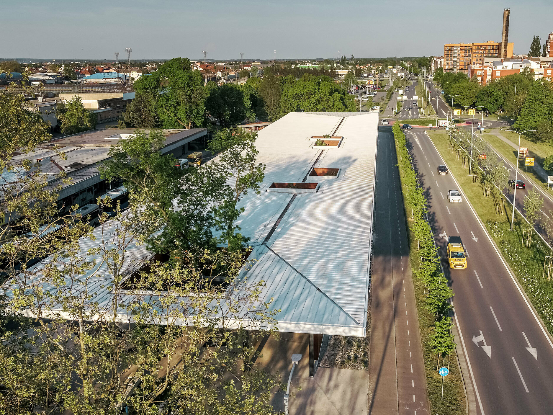 Na obrázku je široký pohled na moderní autobusovou zastávku v Chorvatsku, zastřešenou bílou střechou z PREFALZ od PREFA. Střecha je přerušena několika obdélníkovými otvory, ze kterých vyčnívají stromy. Zastávka je u široké dvouproudé silnice lemované stromy. V pozadí jsou vidět různé budovy a obytné budovy, které charakterizují městské prostředí.