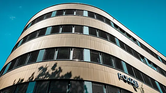 Dramatický pohled zespodu na zakřivenou novostavbu policejní stanice 43 v Hamburku: Výrazná hliníková fasáda PREFA z eloxovaných extrudovaných profilů „Zacke“ ve střední bronzové barvě se stává architektonickým poutačem. Jemně žebrovaný povrch vytváří zářivé světelné odrazy a zdůrazňuje plynulé křivky a horizontální okenní pásy. Díky vysoké odolnosti proti korozi, trvanlivosti a minimální údržbě nabízejí hliníková řešení PREFA udržitelný, funkční a esteticky propracovaný design fasády.