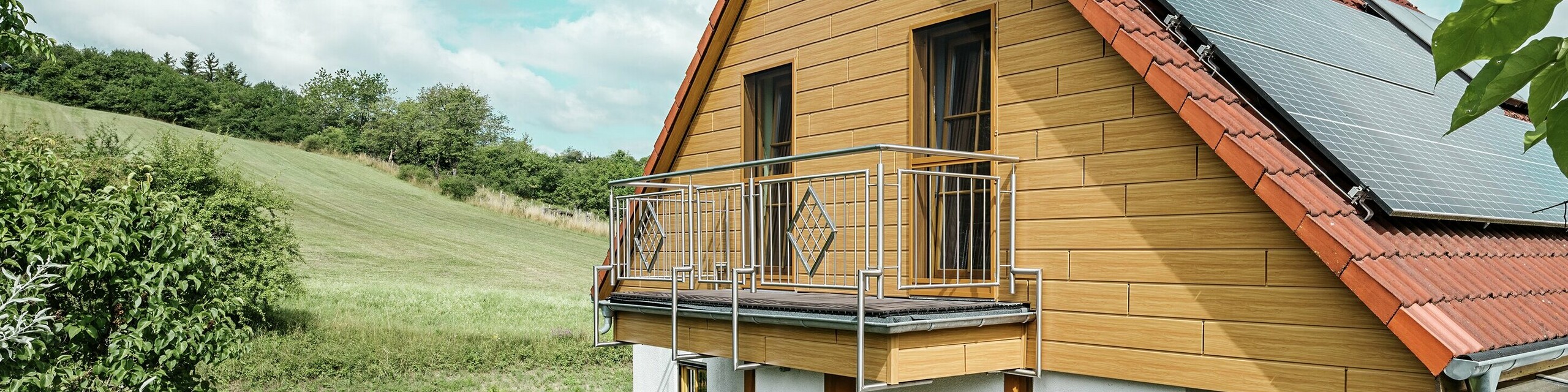 Na této fotografii je zachycen půvabný rodinný dům v Blieskastelu, jehož čerstvě zrekonstruovaný štít s obkladem Siding PREFA v odstínu dub přírodní okamžitě zaujme. Teplý vzhled dřeva z odolného hliníku působivě ladí s červenou taškovou střechou a prostornou zimní zahradou. V obklopení bujné zeleně a pečlivě upravených květinových záhonů vzniká idylická, živá scéna. Stabilní rekonstrukce fasády pomocí produktů PREFA dodává domu nejen přirozený vzhled, ale přesvědčuje také svou odolností vůči povětrnostním vlivům, stálostí barev a dlouhou životností, která prakticky nevyžaduje údržbu.