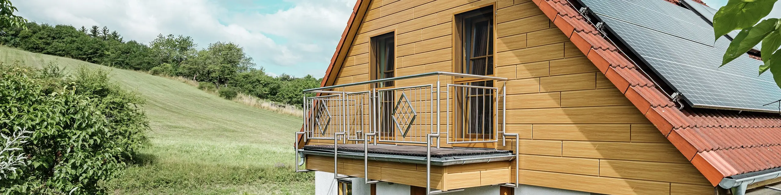 Na této fotografii je zachycen půvabný rodinný dům v Blieskastelu, jehož čerstvě zrekonstruovaný štít s obkladem Siding PREFA v odstínu dub přírodní okamžitě zaujme. Teplý vzhled dřeva z odolného hliníku působivě ladí s červenou taškovou střechou a prostornou zimní zahradou. V obklopení bujné zeleně a pečlivě upravených květinových záhonů vzniká idylická, živá scéna. Stabilní rekonstrukce fasády pomocí produktů PREFA dodává domu nejen přirozený vzhled, ale přesvědčuje také svou odolností vůči povětrnostním vlivům, stálostí barev a dlouhou životností, která prakticky nevyžaduje údržbu.