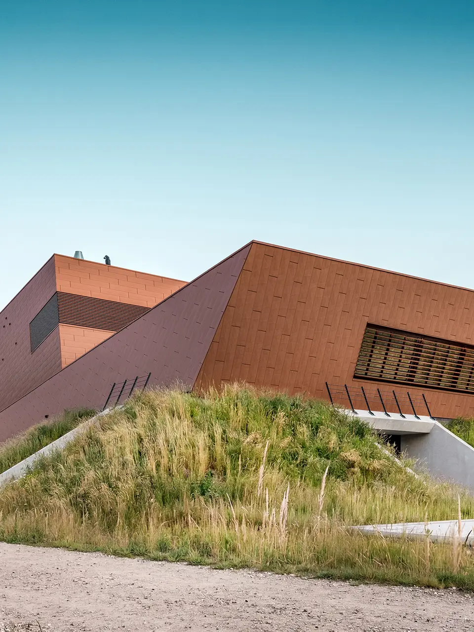 Architektonicky expresivní čerpací stanice pitné vody WML v Lottum se vyznačuje sochařsky skládanými tvary budov, zasazenými do přírodní krajiny nábřeží. Výrazná hliníková fasáda PREFA Siding.X s teplým, rezavějícím vzhledem a fazetovaným, zářivým povrchem je skutečným poutačem a vytváří působivou hru světla a stínu. Horizontální žaluzie a přesné linie zdůrazňují dynamickou geometrii. Lehké, trvanlivé a plně recyklovatelné hliníkové panely PREFA nabízejí maximální odolnost proti povětrnostním vlivům, udržitelnou konstrukci a nenáročný na údržbu pláště budovy.