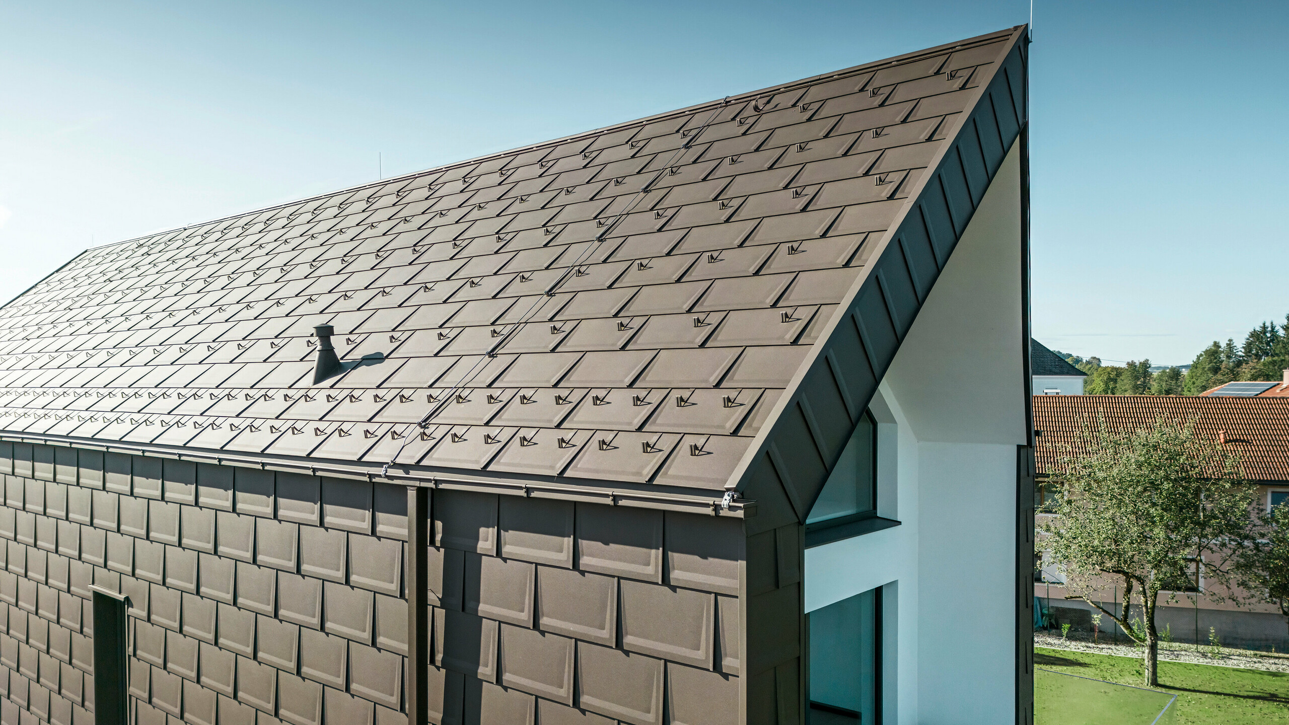 Perspektivní pohled na střechu a fasádu moderního rodinného domu v rakouském Neukirchenu. Střešní panel PREFA R.16 v hnědé barvě P.10 nabízí estetické a funkční zastřešení, které dokonale zapadá do architektonického řešení domu. Ostrý hřeben střechy a přesné uspořádání střešních panelů dokládají kvalitu PREFA. Maloformátové hliníkové výrobky výborně ladí s bílou omítnutou fasádní částí a okolní zelenou krajinou.