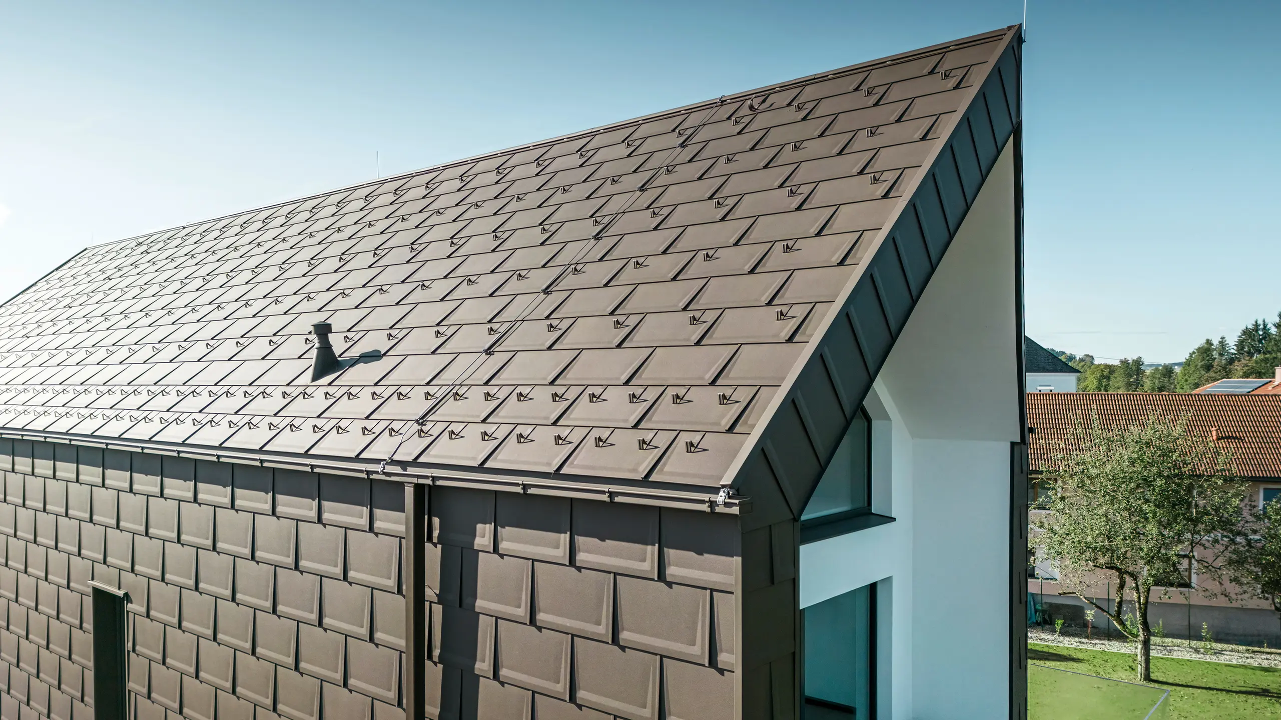 Perspektivní pohled na střechu a fasádu moderního rodinného domu v rakouském Neukirchenu. Střešní panel PREFA R.16 v hnědé barvě P.10 nabízí estetické a funkční zastřešení, které dokonale zapadá do architektonického řešení domu. Ostrý hřeben střechy a přesné uspořádání střešních panelů dokládají kvalitu PREFA. Maloformátové hliníkové výrobky výborně ladí s bílou omítnutou fasádní částí a okolní zelenou krajinou.