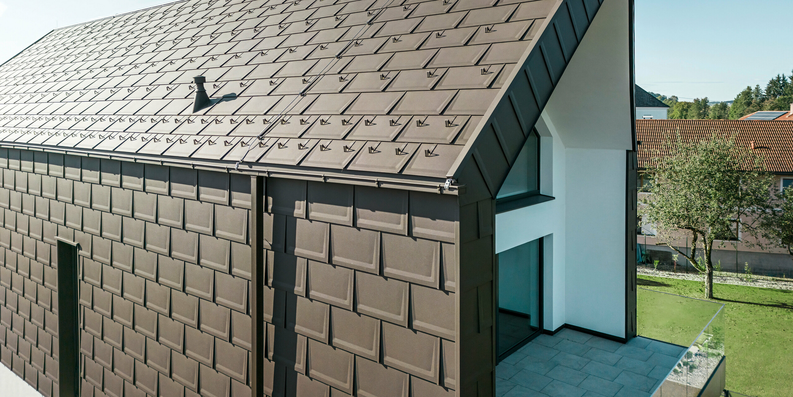 Perspektivní pohled na střechu a fasádu moderního rodinného domu v rakouském Neukirchenu. Střešní panel PREFA R.16 v hnědé barvě P.10 nabízí estetické a funkční zastřešení, které dokonale zapadá do architektonického řešení domu. Ostrý hřeben střechy a přesné uspořádání střešních panelů dokládají kvalitu PREFA. Maloformátové hliníkové výrobky výborně ladí s bílou omítnutou fasádní částí a okolní zelenou krajinou.