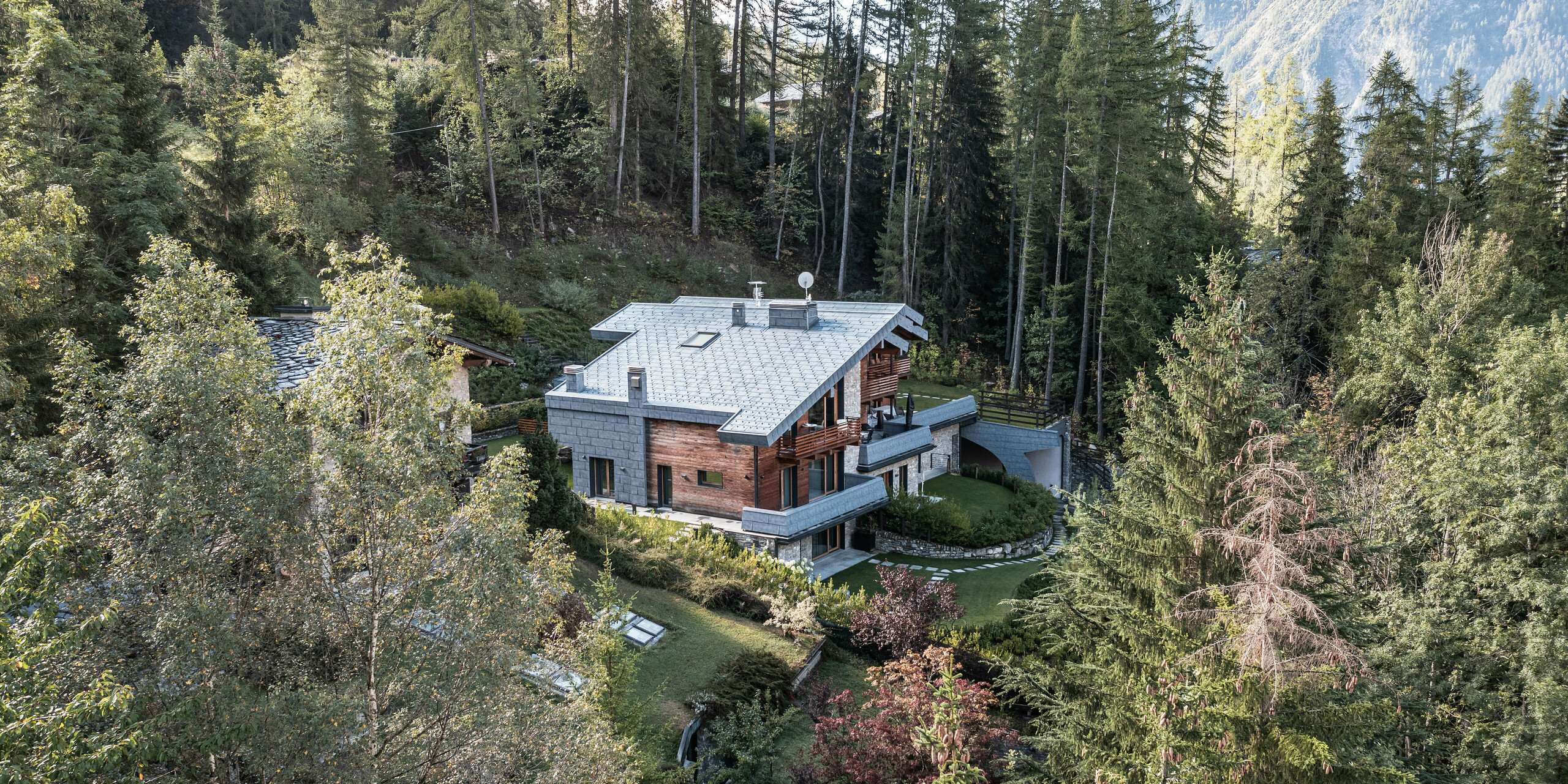 Letecký pohled na impozantní vilu v Courmayeur, obklopenou hustými lesy zvýrazňující přírodní architekturu ze dřeva, kamene a PREFA hliníku. Střešní a fasádní panely FX.12 v břidlicové barvě od PREFA působí jako poutače a poskytují budovu dlouhodobou ochranu. Stupňovité uspořádání stavebních částí umožňuje integraci do strmé krajiny, zatímco několik balkonů a dobře udržovaných zahrad nabízí příjemnou venkovní plochu, která zve k odpočinku v klidném alpském prostředí.