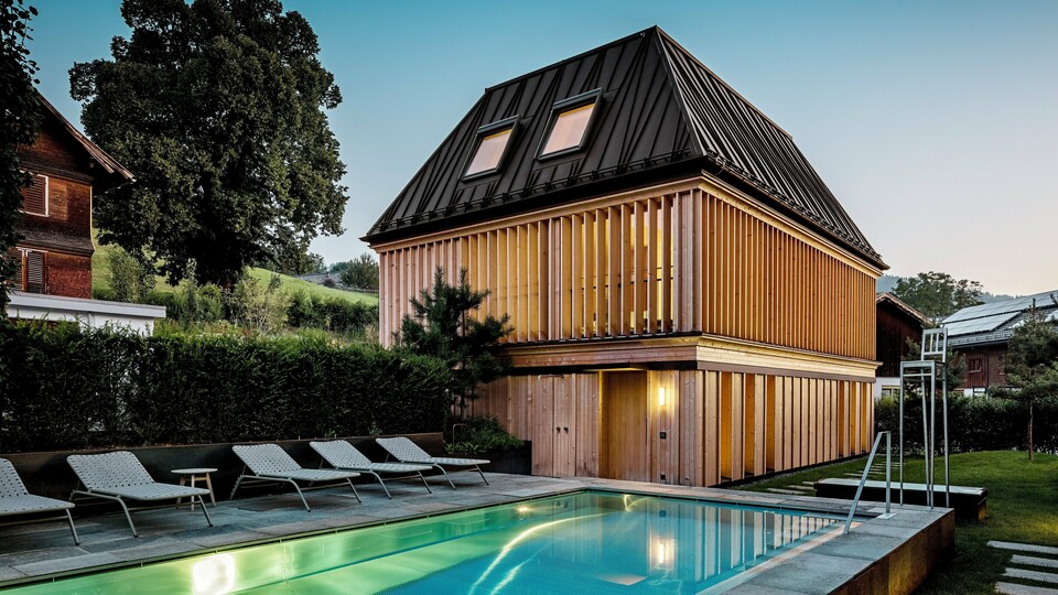 „Lázně“ hotelu Hirschen ve Schwarzenbergu za soumraku s výhledem na osvětlený venkovní bazén v popředí. Budova zaujme moderní valbovou střechou z PREFALZu v oříškově hnědé barvě P.10 s precizně provedenými, částečně zkosenými stojatými drážkami. Dvě střešní okna zajišťují přirozené světlo uvnitř. Fasáda se skládá z vertikálních dřevěných lamel od podlahy až ke stropu, které dodávají budově strukturu a lehkost. Ve spodní části převládá přírodní dřevo. Obklopen upravenou zelení a zasazený do zvlněných kopců Bregenzerwaldu, vytváří harmonickou směs architektury, přírody a relaxace.