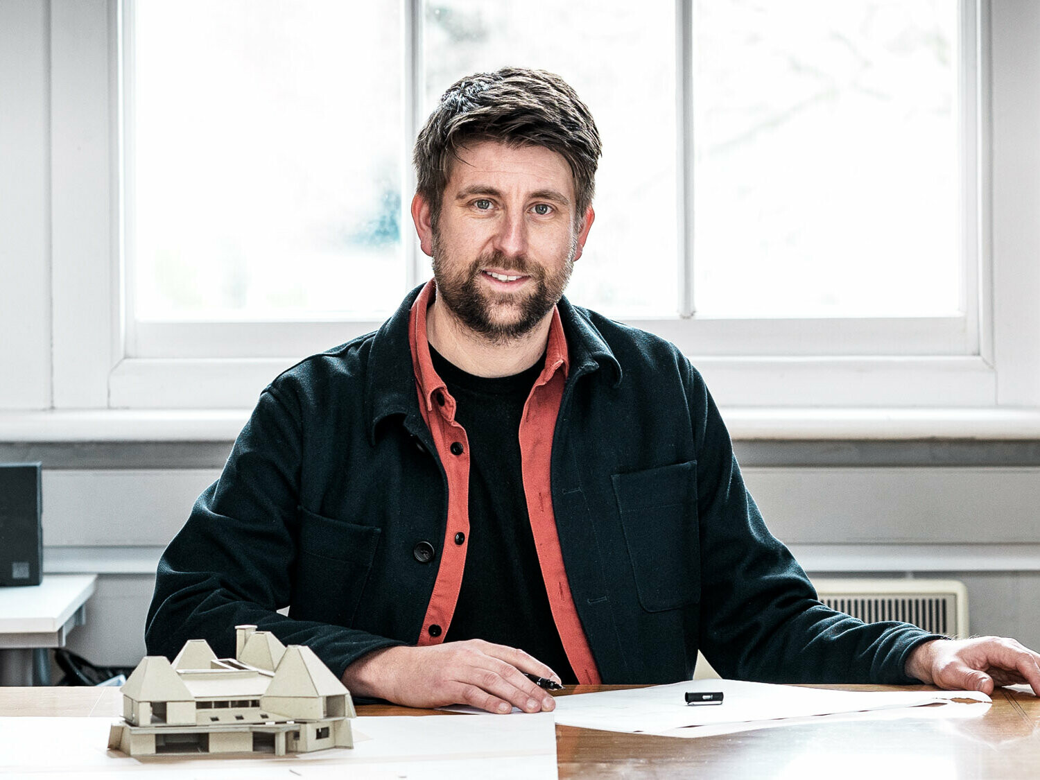Portrét hlavního architekta Tima Howella z britské firmy Mitchell Evans Architects, sedícího u stolu před modelem objektu