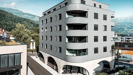 Vnější pohled na Bernerhaus v Buchsu s jeho výrazným trojúhelníkovým tvarem a jemně zaoblenými balkony. Fasáda, obložená PREFA fasádními šindeli v barvě P.10 světle šedé, obaluje budovu jako šupinovitý hliníkový plášť a vytváří precizní a živoucí fasádu. Nad masivním betonovým základem s elegantními arkádami působí obzvláště silně lehká, povětrnostním vlivům odolná hliníková fasáda PREFA. Odolná, nenáročná na údržbu a rozměrově stabilní udává moderní architektonický tón na pozadí alpských hor.