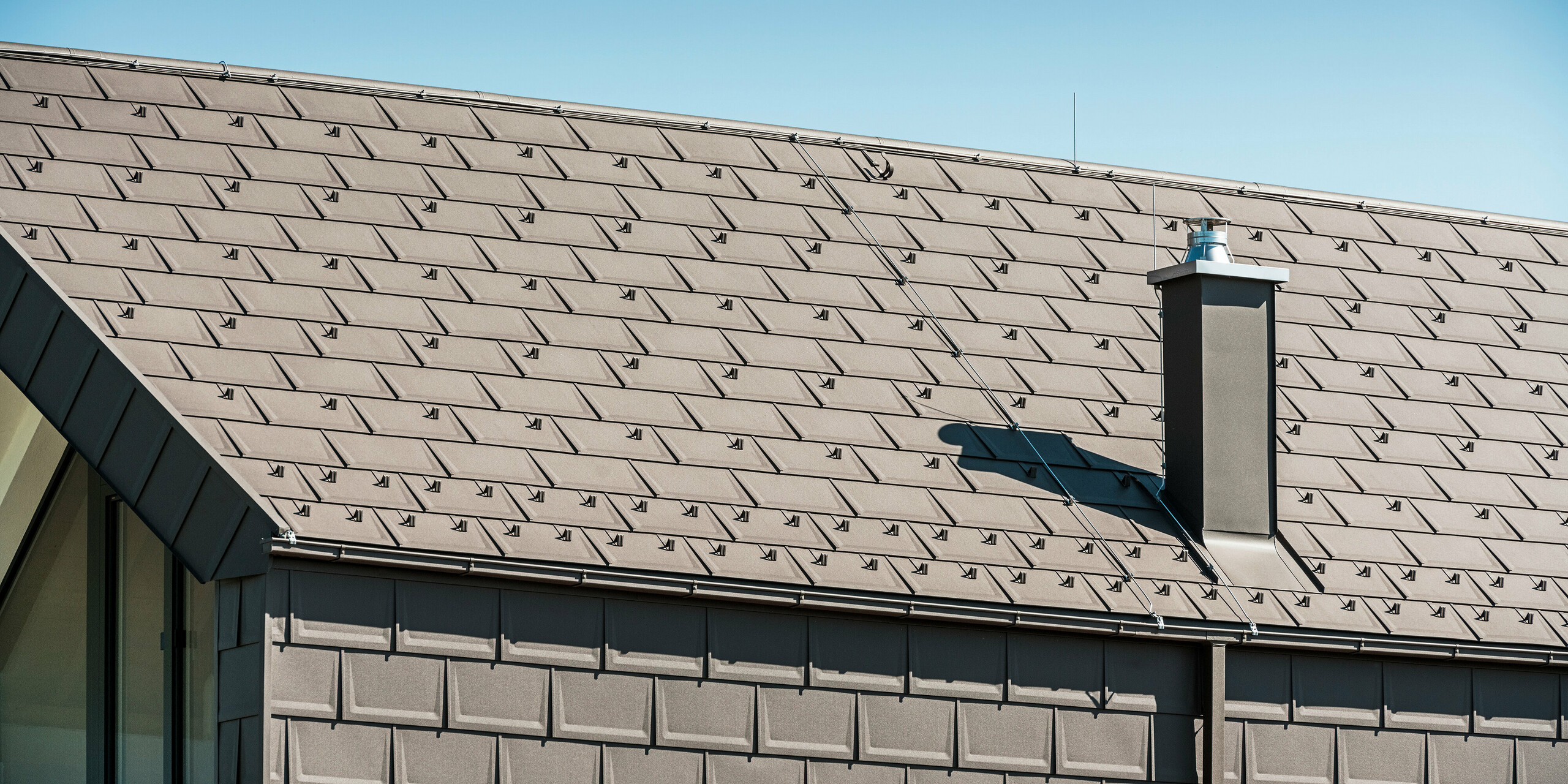 Boční pohled na rodinný dům v rakouském Neukirchenu s precizně položenými střešními panely PREFA R.16 v P.10 hnědé na střeše a fasádě. Na střešní konstrukci je vidět kvalitní řemeslné zpracování a dlouhodobá kvalita výrobků PREFA. Výrazný komín a moderní odvodnění střechy v podobě krabicového žlabu a čtvercového potrubí doplňují střešní systém a zdůrazňují jasné linie a promyšlený design budovy.