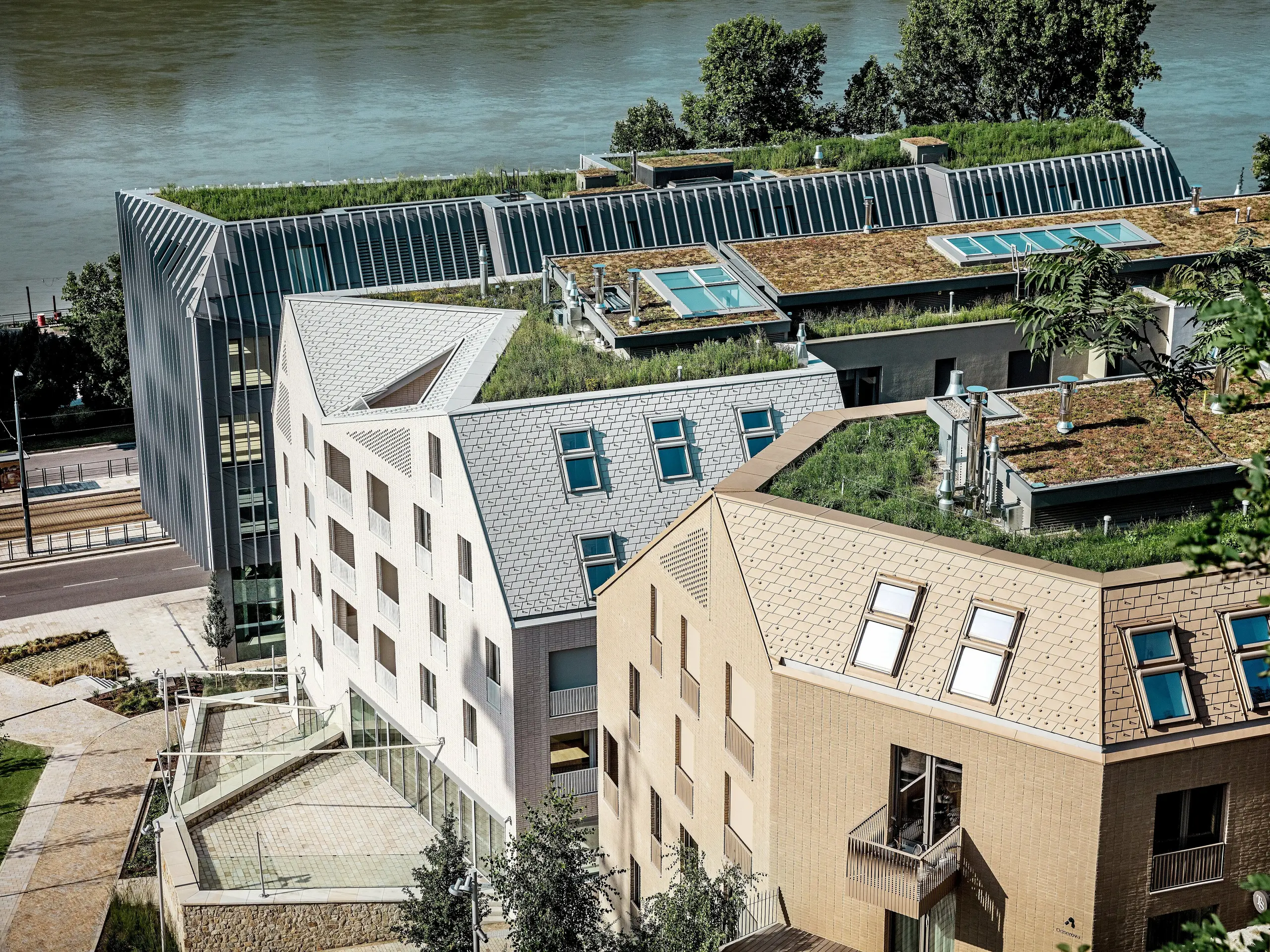 Moderní městská čtvrť v Bratislavě s výraznou architekturou a zelenými střechami přímo na břehu řeky: Jemné, strmě šikmé střechy a vikýře se vyznačují vysoce kvalitními hliníkovými falcovanými šindeli PREFA v odstínech prefa bílá, písková a bronzová, které okamžitě upoutají pozornost. Velkorysá střešní okna, čisté linie a bujná střešní vegetace spojují design a udržitelnost. Odolná a nenáročná hliníková řešení PREFA přesvědčují svou odolností, lehkostí a odolností vůči povětrnostním vlivům – ideální pro náročné městské stavební projekty.