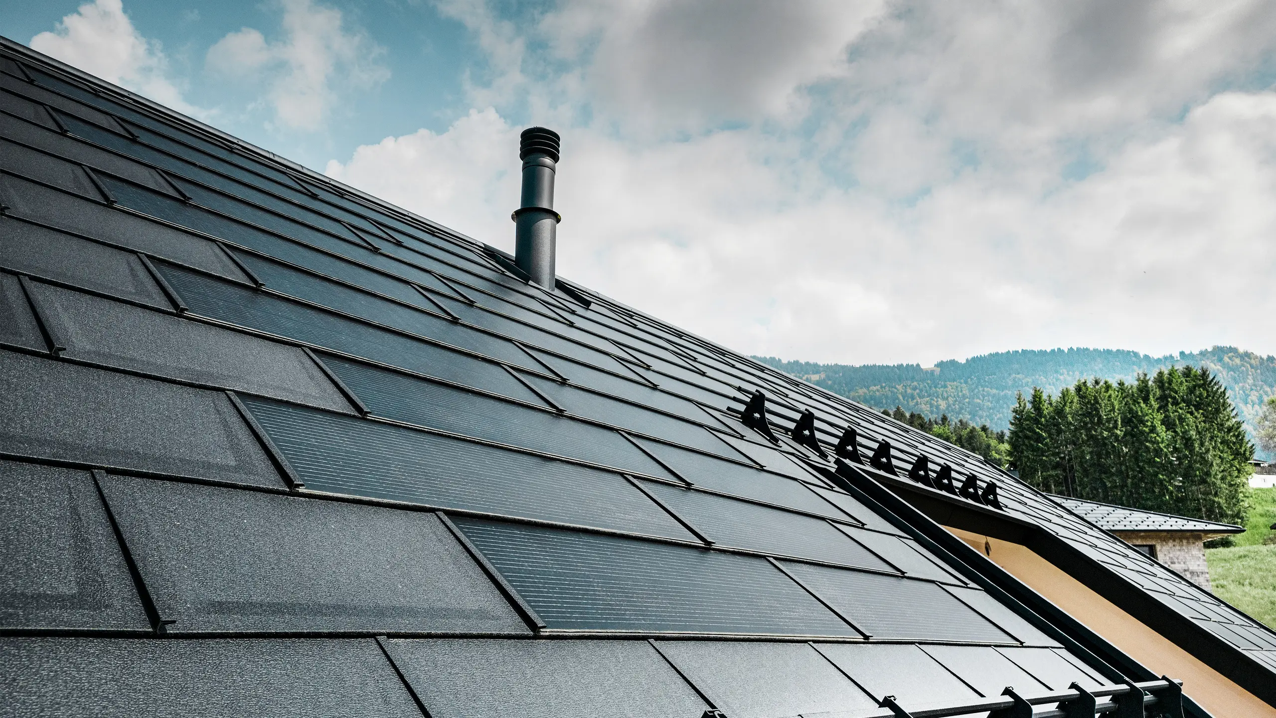 Detailní pohled na solární střešní panel PREFA v kombinaci s osvědčeným střešním panelem PREFA R.16 v barvě P.10 black na ekologickém domě v rakouském Eggu. Integrované fotovoltaické moduly demonstrují bezproblémové spojení obnovitelné energie a důmyslného designu střechy. Černé střešní panely R.16 se systémem ochrany proti sněhu dotvářejí estetický povrch střechy a zároveň zajišťují bezpečnost.