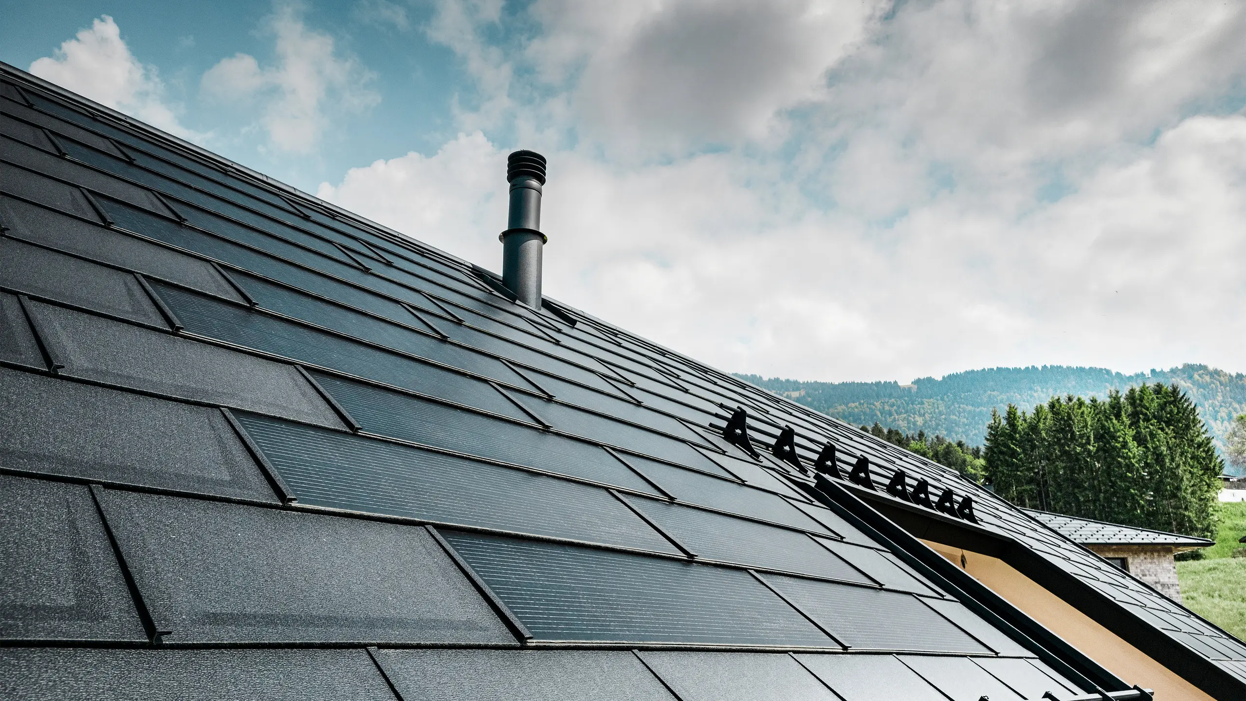 Detailní pohled na solární střešní panel PREFA v kombinaci s osvědčeným střešním panelem PREFA R.16 v barvě P.10 black na ekologickém domě v rakouském Eggu. Integrované fotovoltaické moduly demonstrují bezproblémové spojení obnovitelné energie a důmyslného designu střechy. Černé střešní panely R.16 se systémem ochrany proti sněhu dotvářejí estetický povrch střechy a zároveň zajišťují bezpečnost.
