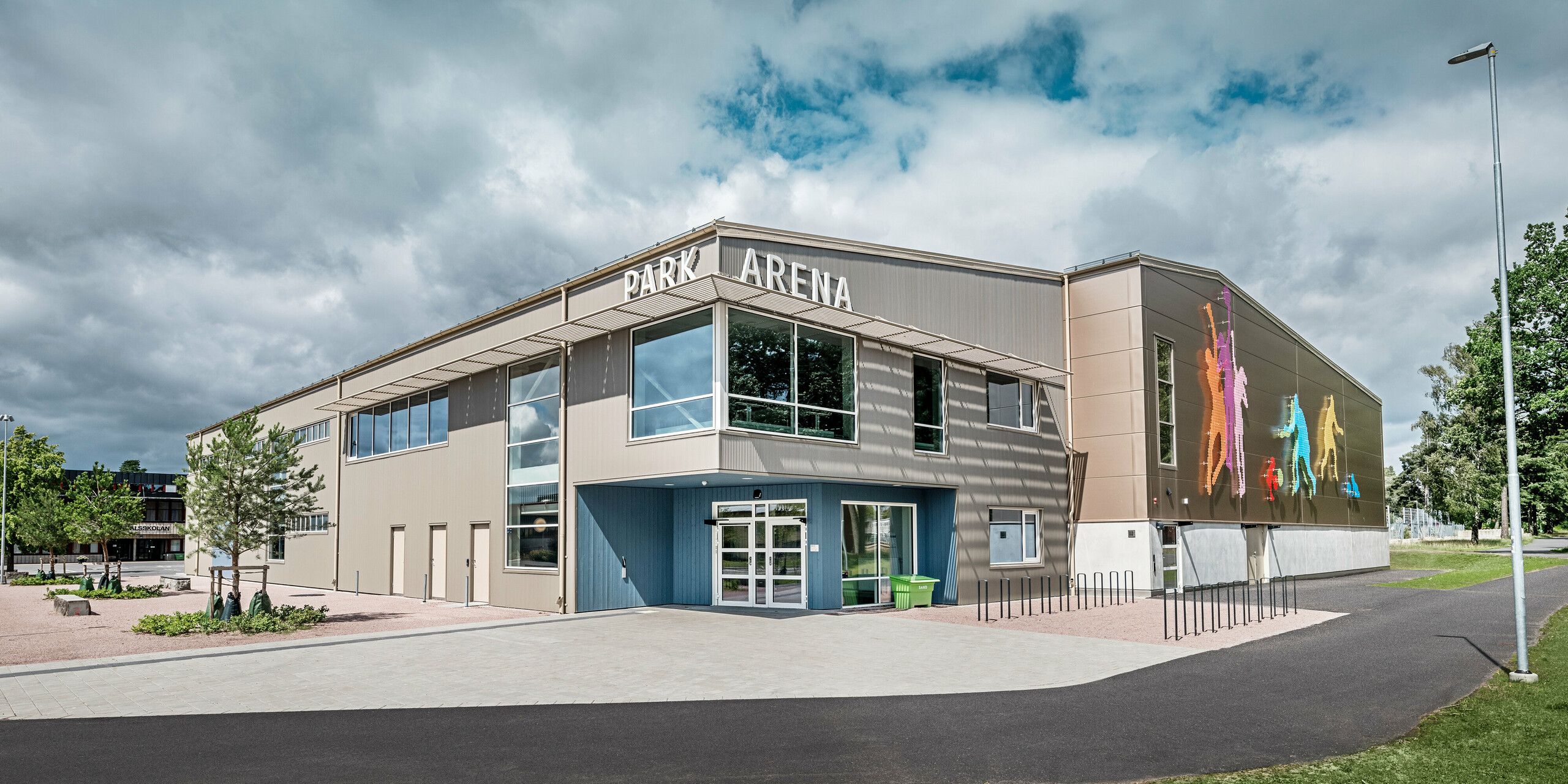 Čelní pohled na Park Arenu ve Värnamo, Švédsko, s moderní fasádou PREFA z pilových profilů v bronzu. Hliníková fasáda nabízí dlouhodobé a odolné řešení, které vyniká výraznými světelnými a stínovými efekty. Umělecky navržená nástěnná grafická instalace vpravo zobrazuje barevné sportovní postavy, které zdůrazňují dynamický charakter sportovní haly. Velká okna propouštějí do budovy dostatek přirozeného světla a posilují moderní design arény.