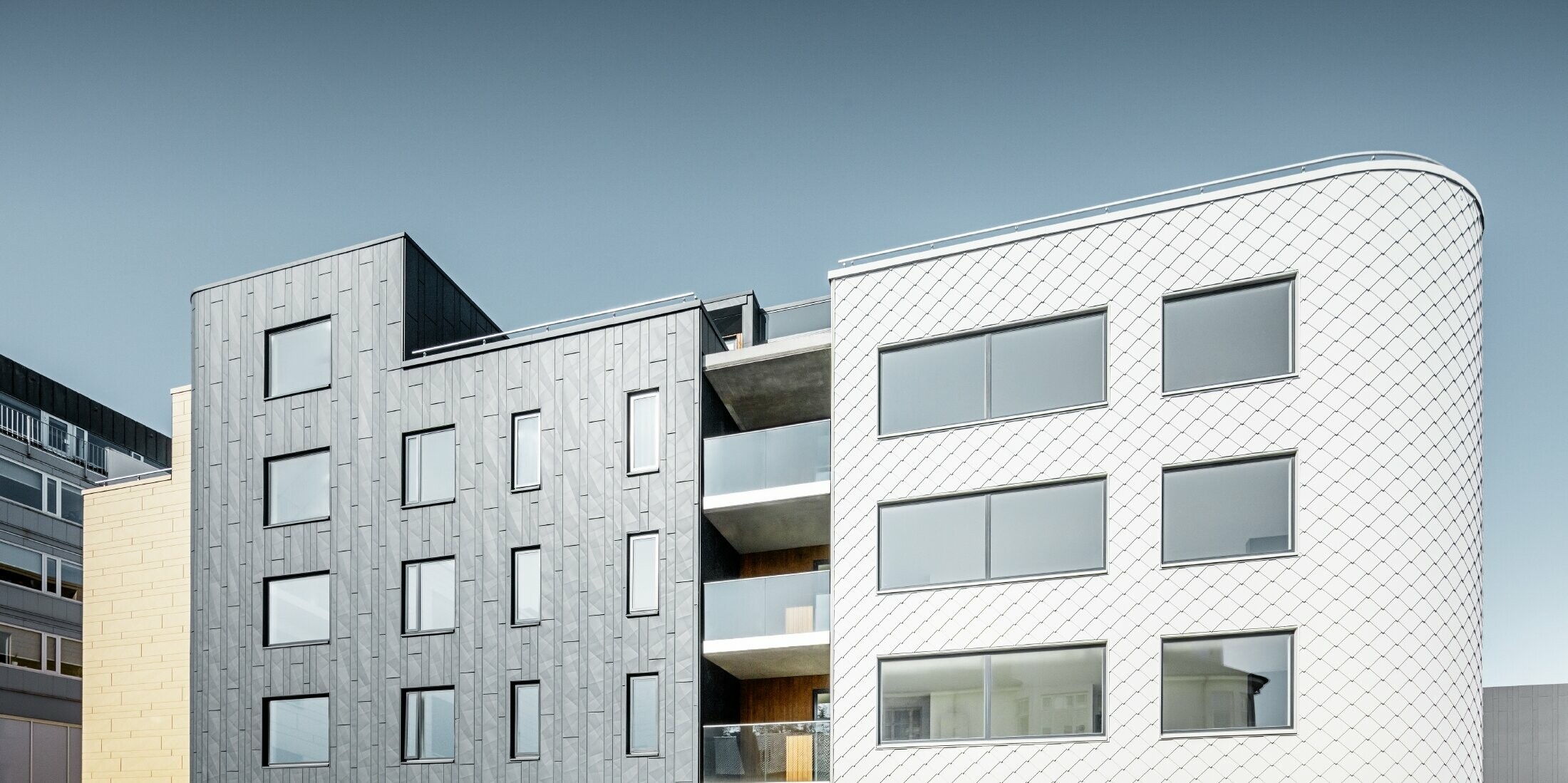 Vertikálně namontované fasádní lamely Siding.X v barvě antracitové na jedné budově, zatímco druhá je pokryta světlými fasádními šablonami 29 x 29
