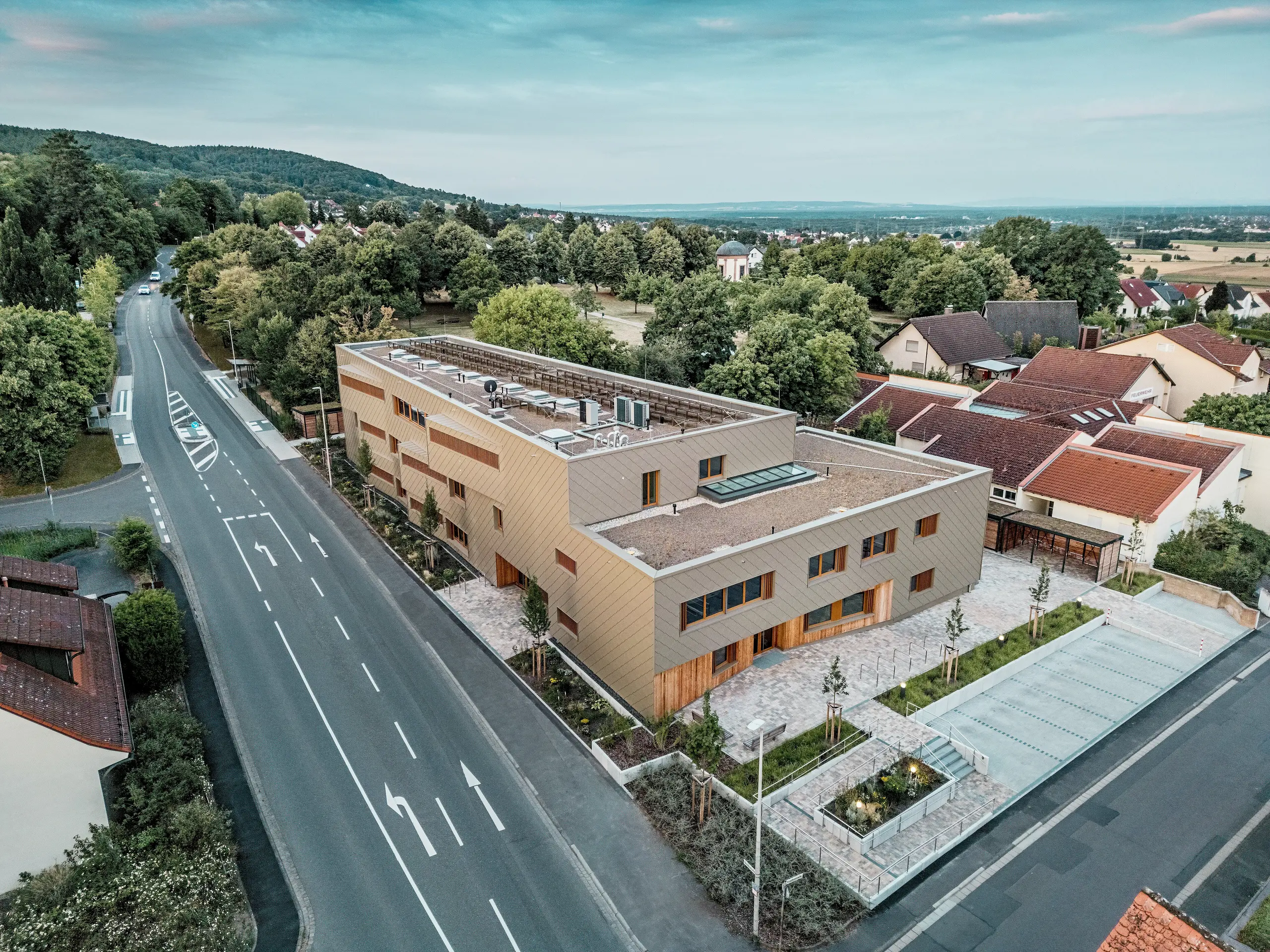 Působivý celkový design městské denní péče v Alzenau je jasně viditelný na leteckém snímku. Hranatá hybridní budova je harmonicky začleněna do klidné obytné čtvrti na okraji lesa Spessart. Fasáda z bronzově zbarvených stěnových panelů PREFA (44 x 44 cm) dodává budově výrazný, vysoce kvalitní vzhled a zdůrazňuje její moderní architekturu. Zelené střechy, prostorné venkovní plochy a dobře navržený dopravní tok podtrhují celkovou udržitelnou koncepci. Kombinace odolného hliníku, dřeva a recyklovaného betonu splňuje nejvyšší ekologické standardy. Tato perspektiva zdůrazňuje promyšlenou integraci do města a multifunkční charakter budovy – průkopnického projektu pro vzdělávání, bydlení a udržitelnost.