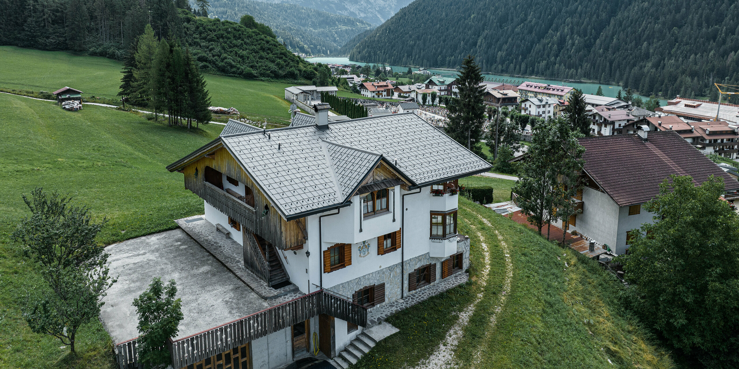 Letecký pohled na půvabnou horskou chatu v italském Auronzo di Cadore po renovaci střechy se hliníkovými taškami PREFA ve světle šedé barvě. Chata se tyčí na zelené louce s působivým pohořím v pozadí a ukazuje malebnou souhru modernizovaných stavebních prvků a tradiční alpské architektury. Nad vstupním prostorem je vidět hrotitý vikýř s dřevěným štítem.