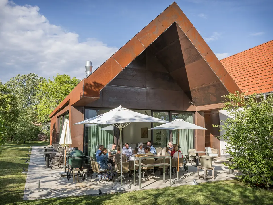 PREFARENZEN 2027 Expertendialog auf der Terrasse einer modernen Winzerresidenz: Jury-Mitglieder sitzen um einen großen Tisch, diskutieren intensiv über Architekturprojekte und blättern in Architekturbüchern. Die Szene wird von natürlichem Licht und grünem Garten umrahmt, während PREFA Aluminiumprodukte an Dach und Fassade des Gebäudes sichtbar sind. Das Bild vermittelt kreative Auseinandersetzung, konzentrierte Analyse und lebendigen Austausch. PREFA Dächer und Fassaden stehen für innovative, langlebige und ästhetisch anspruchsvolle Lösungen, die Design, Funktion und Inspiration in Architekturprojekten verbinden.
