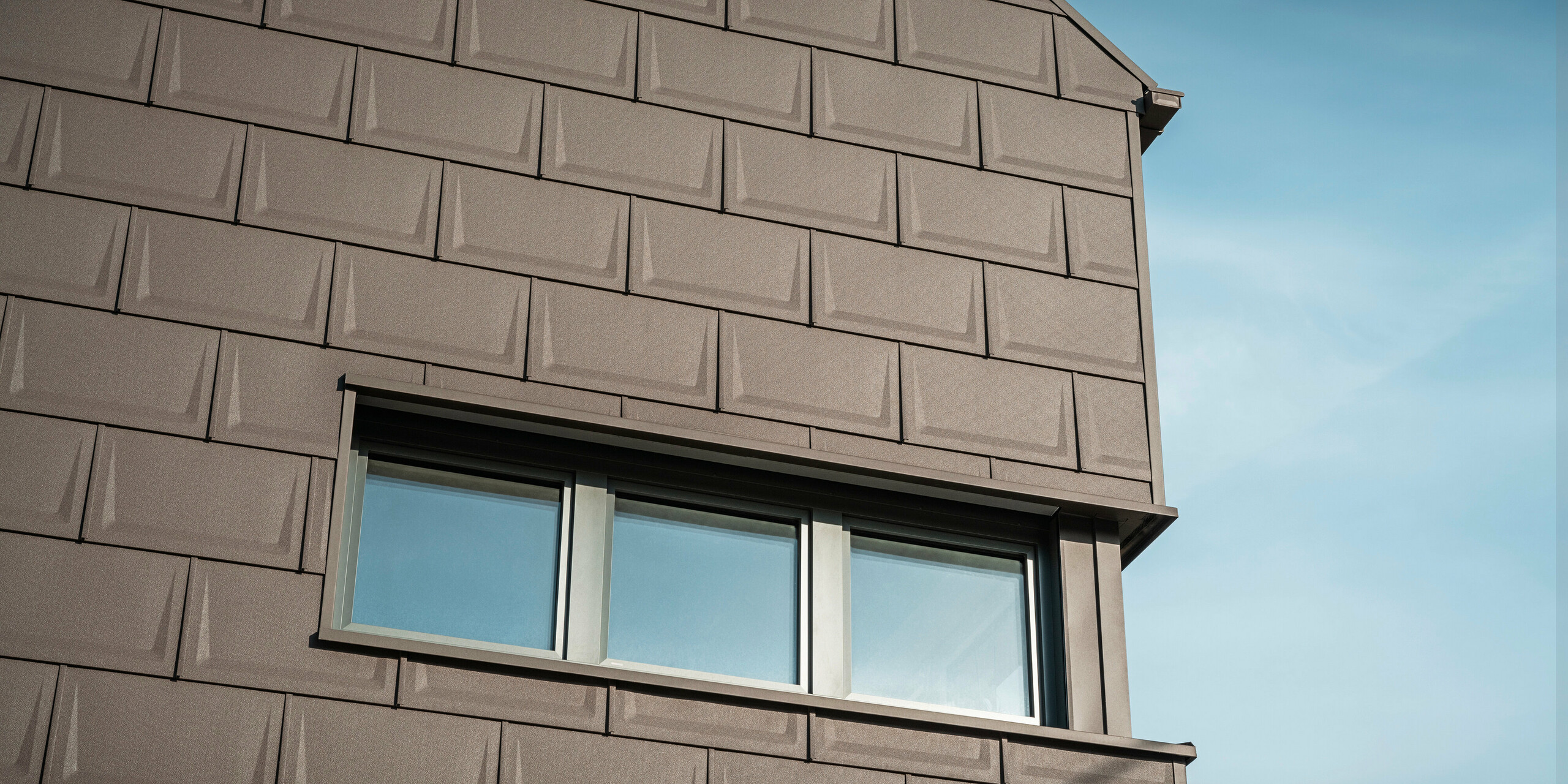 Detailní záběr na fasádu rodinného domu v Neukirchen, Rakousko, navržený s inovativním střešním panelem PREFA R.16 v hnědé P.10, který se používá jako speciální objektové řešení pro střechu i fasádu návrh horního patra. Tato jedinečná aplikace zajišťuje harmonický a estetický vzhled a podtrhuje všestrannost hliníkových výrobků PREFA. Jednotné barevné řešení a promyšlený designový koncept budovy zdůrazňují všestranné designové možnosti PREFA.