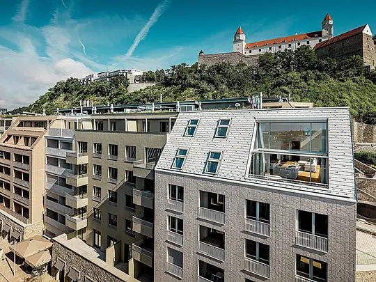 Moderne Dachlandschaft im Stadtquartier Vydrica in Bratislava mit Blick zur Burganlage: Steil geneigte Dachflächen mit großzügigen Fensteröffnungen und klaren Linien werden von PREFA Aluminium Dachschindeln in Prefaweiß und Sandbraun elegant in Szene gesetzt. Die präzise Verarbeitung und die harmonische Verbindung von Dach und Fassade schaffen einen starken visuellen Eyecatcher über den Dächern der Stadt. PREFA Aluminium überzeugt durch Leichtigkeit, hohe Witterungsbeständigkeit und langlebige Qualität – ideal für komplexe, architektonisch anspruchsvolle Wohnbauten.