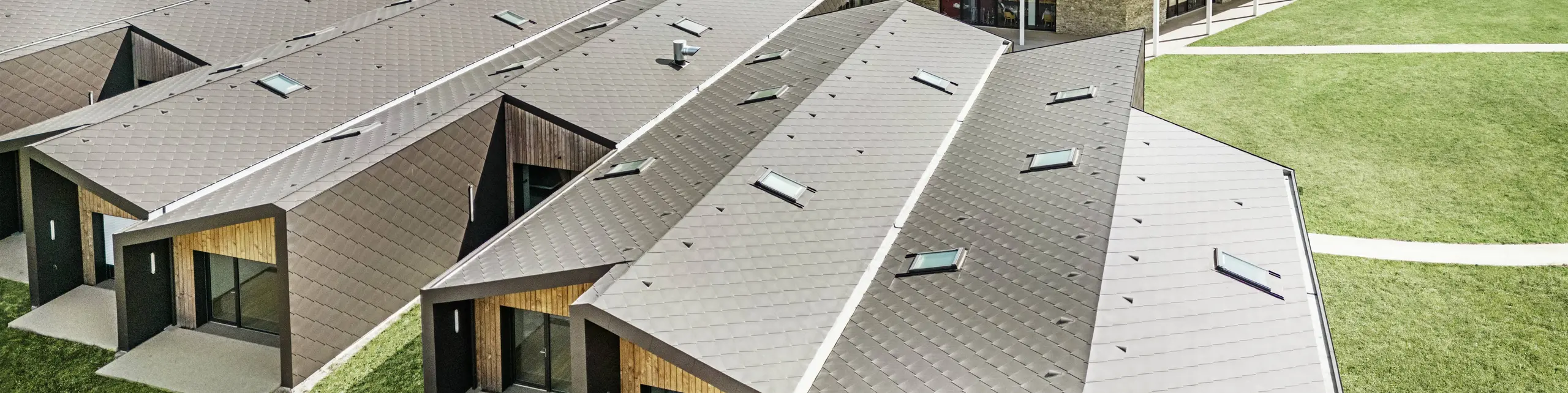 Na obrázku je letecký pohled na moderní komplex budov opláštěný falcovanými a fasádními šablonami PREFA 44 × 44 v barvě P.10 oříškové. Střešní krajina se vyznačuje geometrickými tvary, které jsou kombinovány s velkými prosklenými čely a dřevěnými prvky. V pozadí je vidět starý kostel, který tvoří harmonický kontrast k moderní architektuře. Zelený trávník a okolní příroda podtrhují otevřený a udržitelný design celého areálu.