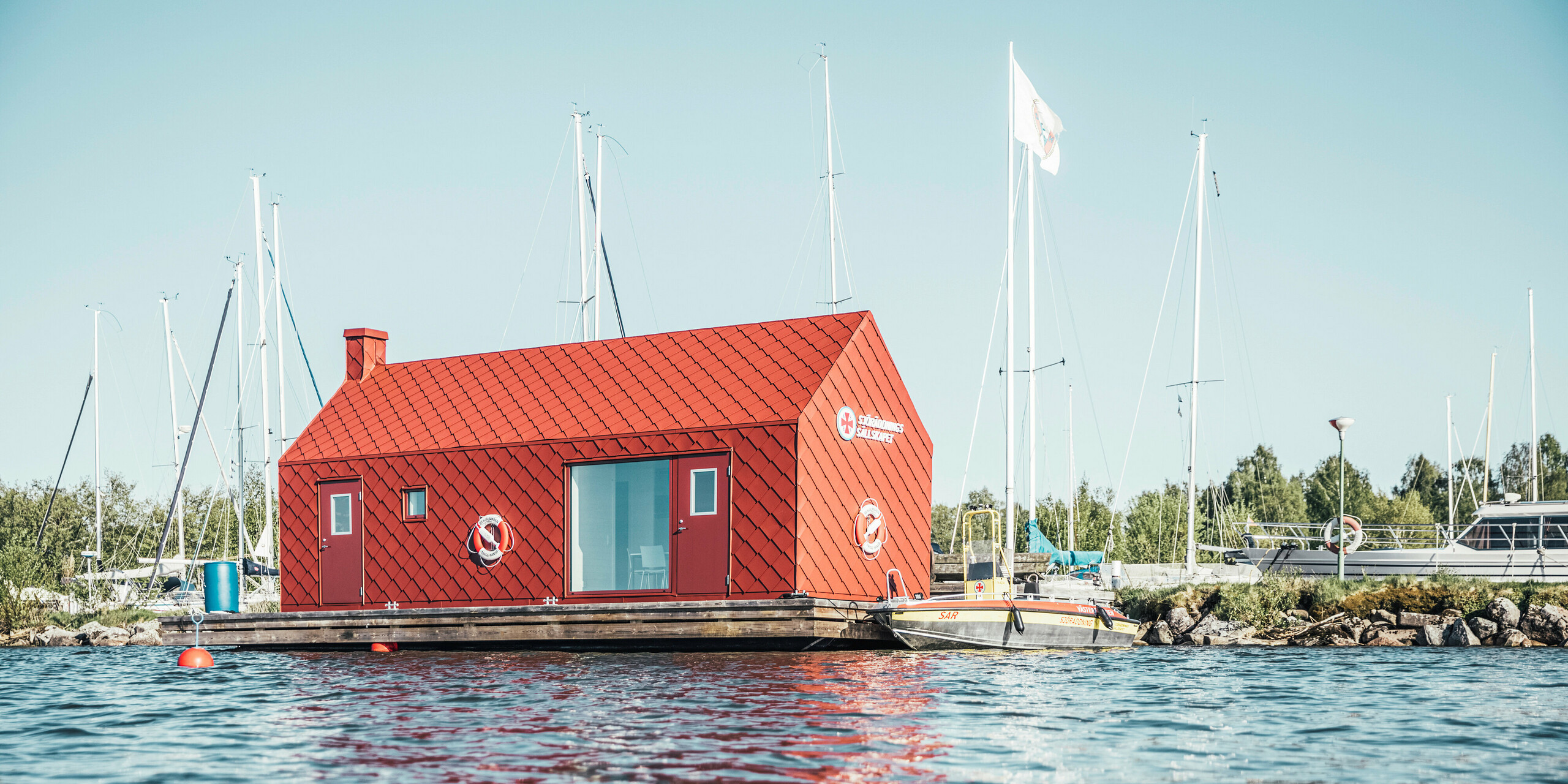 Plovoucí staniční dům vodních záchranářů na břehu jezera Hjälmaren obklopený klidnými vodami a stěžněmi plachetnic září ve švédském slunci. Živý, tmavě červený hliníkový plášť vyrobený z PREFA falcovaných a fasádních šablon 29×29 poskytuje nejen vizuální teplo, ale také strukturální integritu, aby odolal drsným klimatickým podmínkám. Tato budova, vybavená záchrannými kruhy a umístěná jako strážce bezpečnosti, symbolizuje spolehlivost a připravenost vodních záchranářů, kterým slouží jako domov.