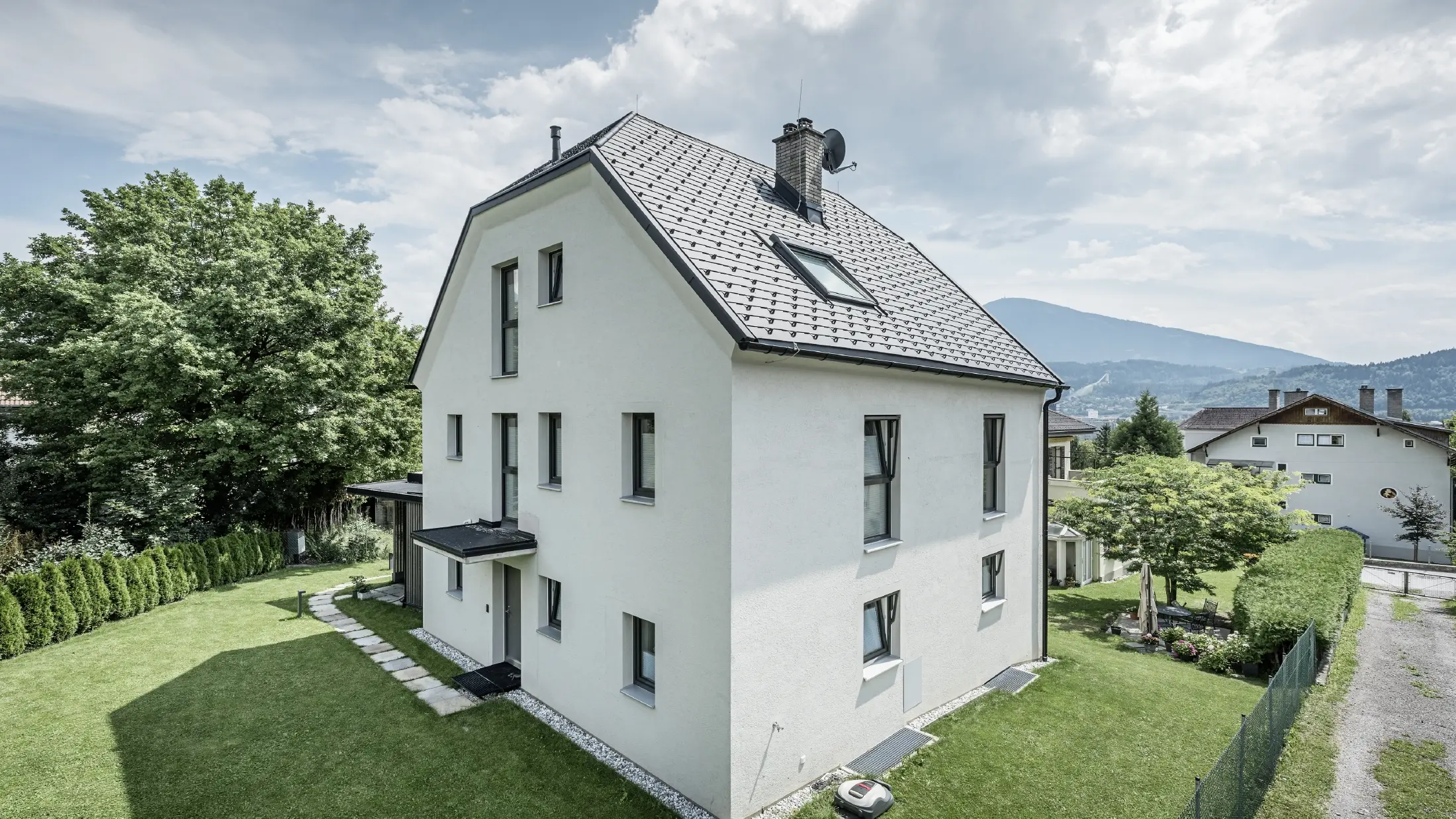 Moderní třípodlažní rodinný dům se světlou omítkovou fasádou a tmavě šedou sedlovou střechou z hliníku od PREFA. Budova má několik úzkých oken a antracitovou odvodnění střechy. Stojí v upravené zahradě s trávníkem, stromy a živými ploty, v pozadí jsou vidět další obytné domy a hornatá krajina.