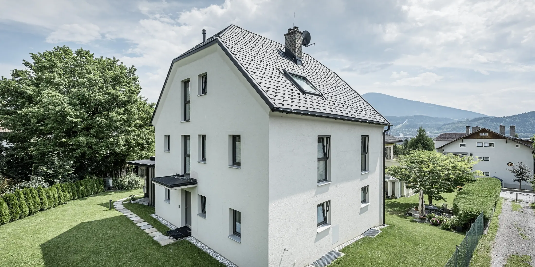 Moderní třípodlažní rodinný dům se světlou omítkovou fasádou a tmavě šedou sedlovou střechou z hliníku od PREFA. Budova má několik úzkých oken a antracitovou odvodnění střechy. Stojí v upravené zahradě s trávníkem, stromy a živými ploty, v pozadí jsou vidět další obytné domy a hornatá krajina.