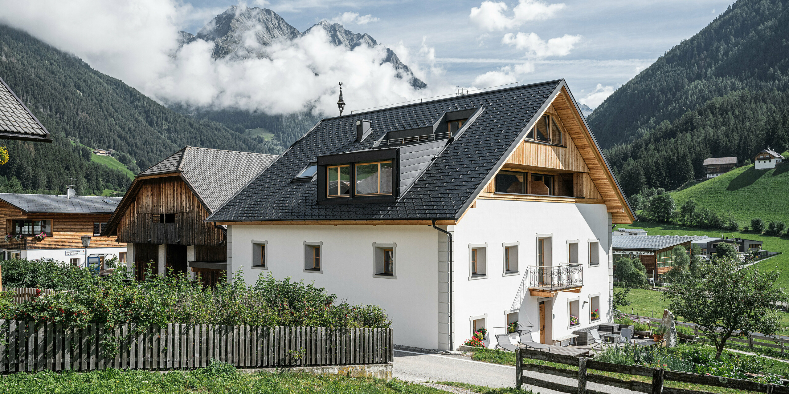 Úplný výhled na prázdninové apartmány Ausserwegerhof v idylickém údolí Antholz, Jižní Tyrolsko, pokryté střešním šindelem PREFA DS.19 v antracitové barvě. Tradiční stavba ladí s moderními prvky a nabízí malebný výhled na majestátní horské pozadí, tajemně obklopené mraky. Ausserwegerhof uprostřed obrázku je obklopen klasickými farmami a zelenými loukami.