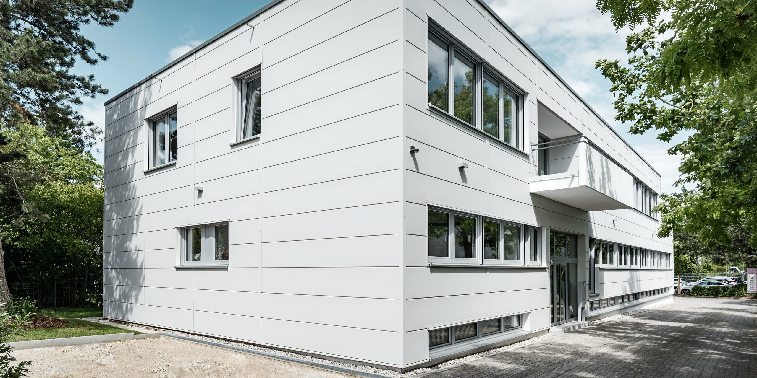 Boční pohled na budovu se zavěšenou provětrávanou fasádou od PREFA v barvě P.10 prefa bílé. Na obrázku vidíme hlavní vchod do budovy.