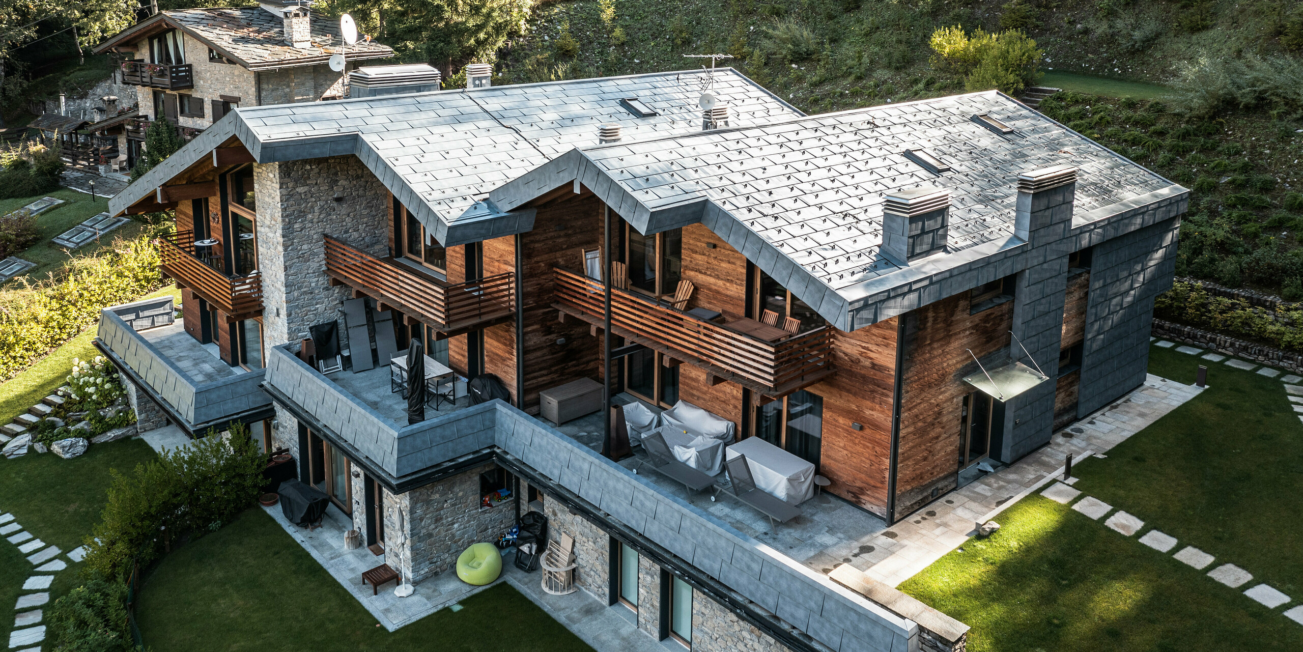 Letecký pohled na prostornou vilu v Courmayeur, zasazenou do přírodní lesní krajiny. Architektura kombinuje dřevo, kámen a moderní hliníkové panely PREFA FX.12 v barvě P.10 břidlicové. Charakteristické balkony a terasy nabízejí rozsáhlé venkovní plochy pro relaxaci. Design zahrady s přesnými zelenými plochami a kamennými prvky ladí s rustikální a moderní strukturou vily a nabízí úchvatné výhledy na Alpy.