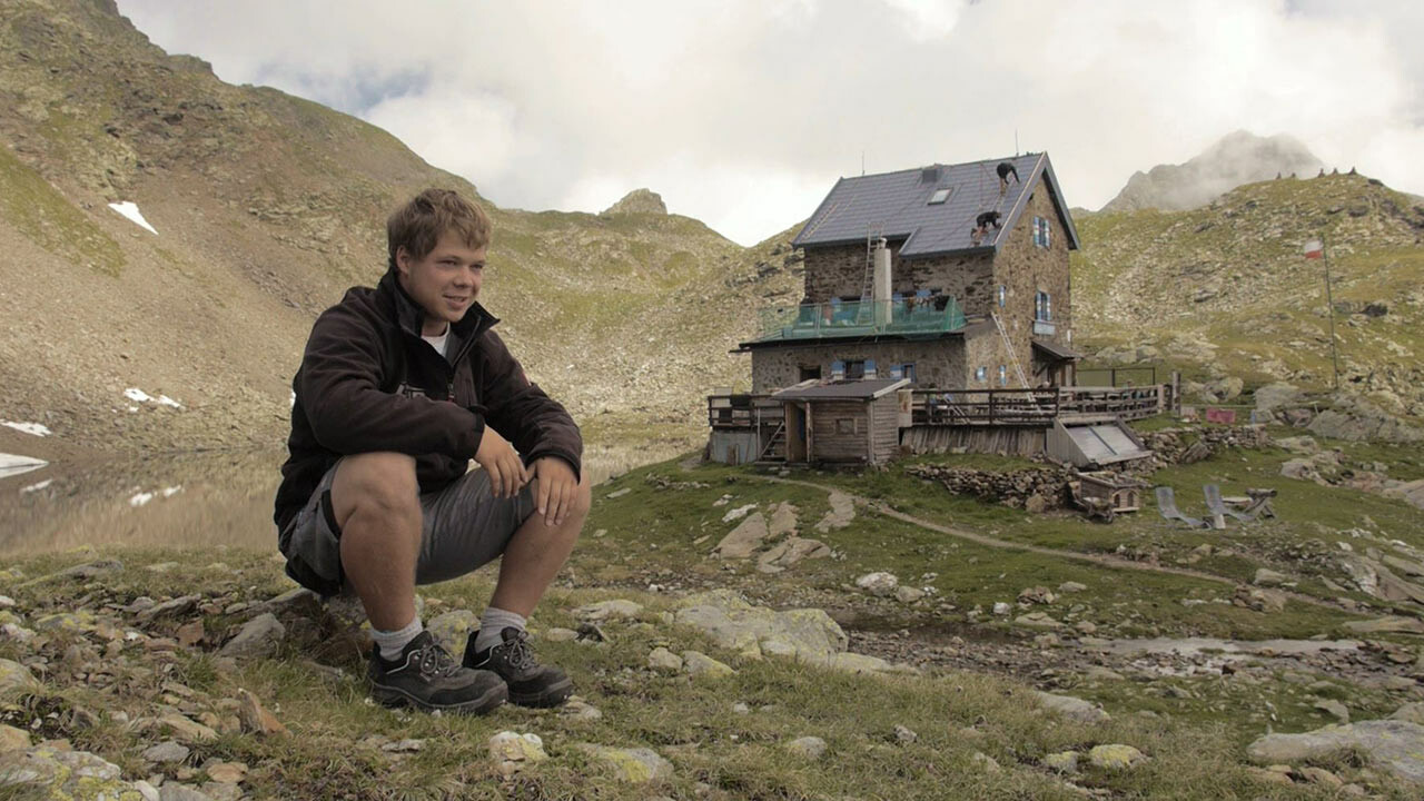 Učeň Jakob Schöttl sedí na kameni. V pozadí je vidět chata Flagger Schartenhütte s novou střešní krytinou z panelů PREFA.