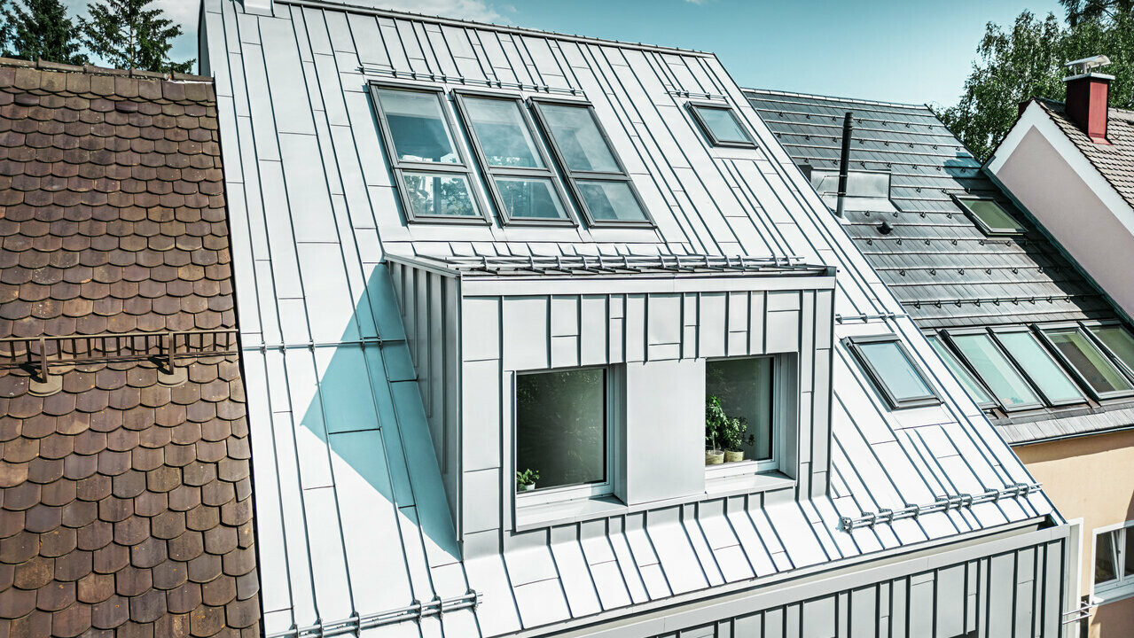 Moderní řadový dům po rekonstrukci střechy a fasády s hliníkovými výrobky PREFA. Střechu tvoří hliníkové pásy v barvě stříbrné metalízy, které mají texturovaný, matný povrch a odrážejí světlo. Fasáda domu je také obložena vertikálními hliníkovými prvky, které poskytují kontrast k tradičním, terakotově zbarveným taškovým střechám přilehlých domů.
