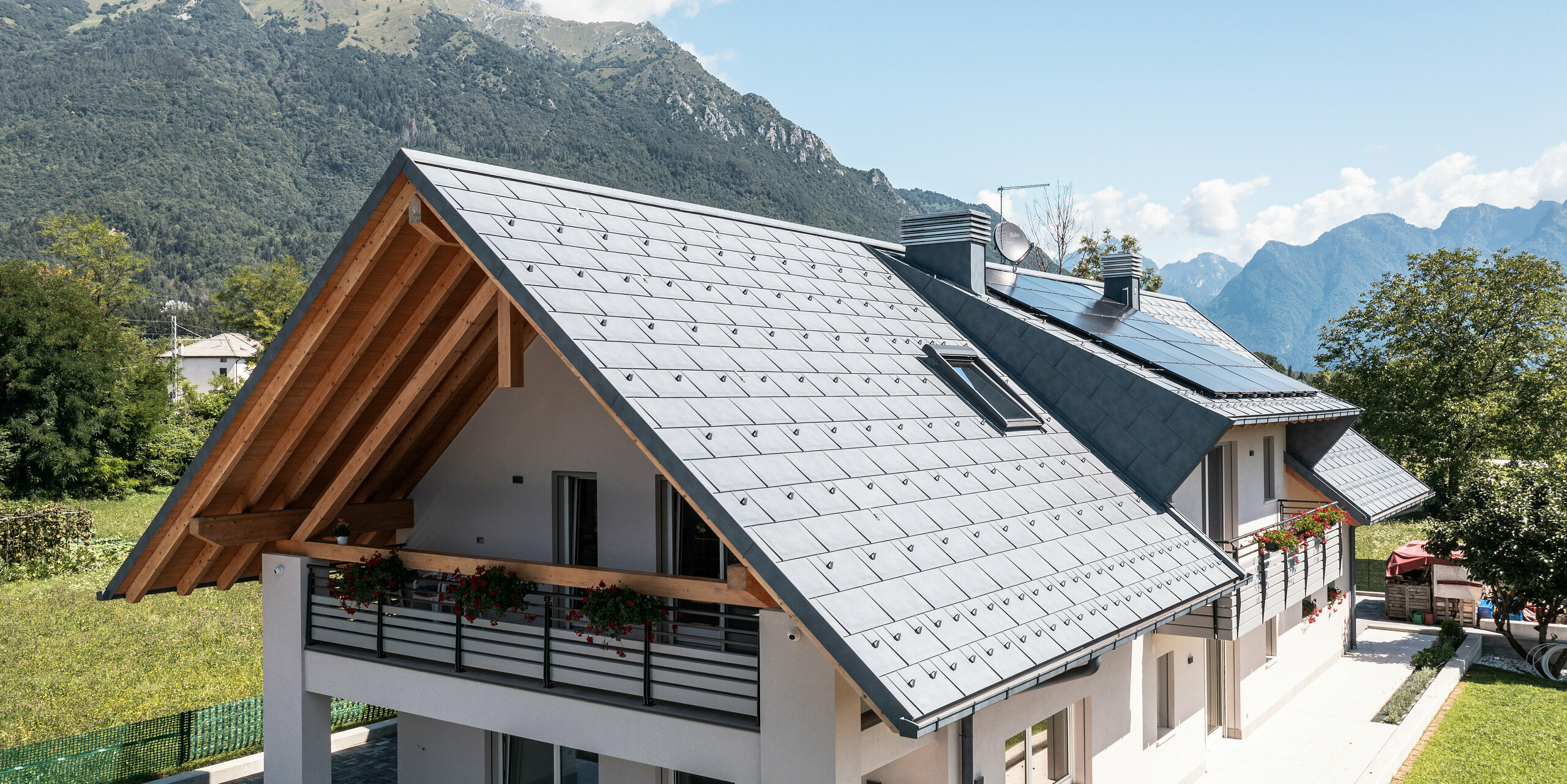 Idylický rodinný dům v Bellunu vybavený střešními panely PREFA R.16 v antracitové barvě, který stojí uprostřed svěží zelené louky. Čisté linie architektury a moderní střecha vytvářejí nádherný kontrast k tradičním venkovským domům v pozadí a okolní horské krajině.