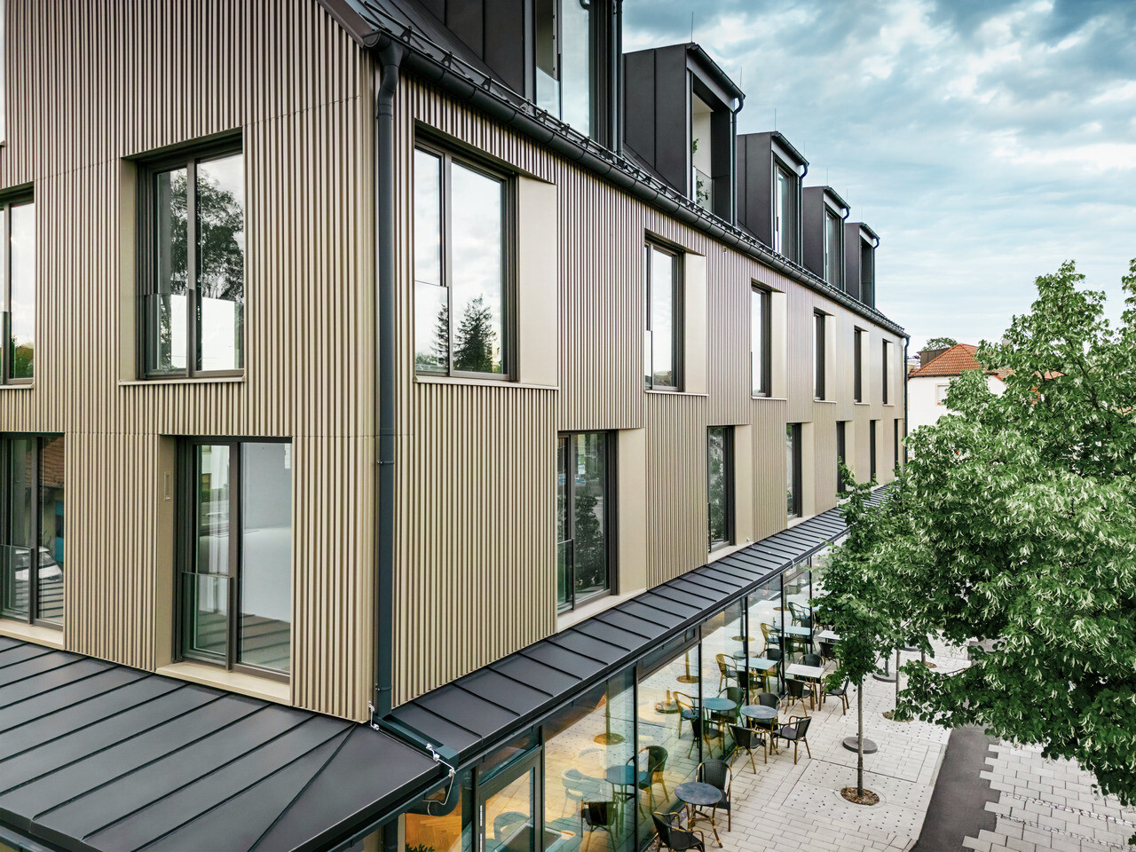 Na obrázku je moderní budova ve Feldkirchu, kterou navrhl architekt Stefan Heigl. Fasáda je vyrobena z Prefalz v barvě bronz a sedlová střecha je pokryta Prefalz v černo-šedé. Velká okna poskytují dostatek světla uvnitř, zatímco prosklená část přízemí nabízí příjemný jídelní kout. Výběr kvalitních materiálů a elegantní design dodávají budově moderní a stylový vzhled.