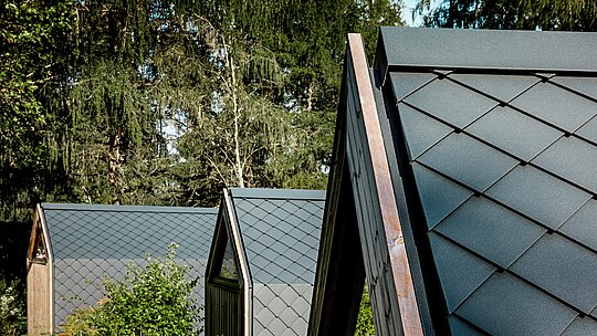 Několik minimalistických domků ve tvaru písmene A v Tajné zahradě koi je harmonicky uspořádáno na okraji lesa u jezera Natterer. Střechy jsou pokryty malými hliníkovými šablonami PREFA ve tvaru diamantu v antracitové barvě P.10, které vytvářejí výraznou texturu připomínající rybí šupinky. Matný povrch působí klidně a elegantně, zatímco odolný a povětrnostním vlivům odolný hliník PREFA nabízí optimální ochranu v alpském prostředí a dokonale propojuje design s přírodou.